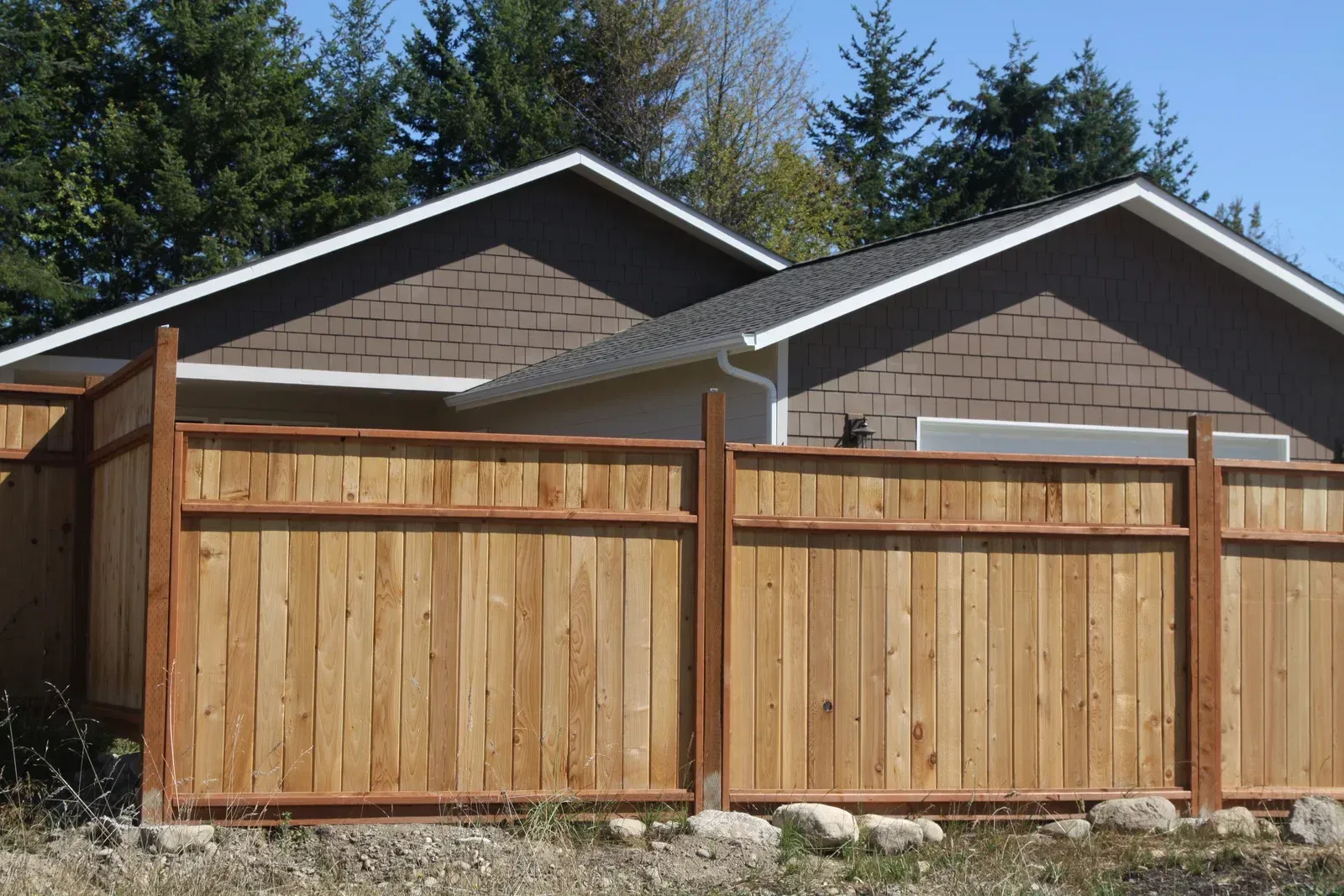 Wooden fence in front of a house with a brown roof and trees in the background.