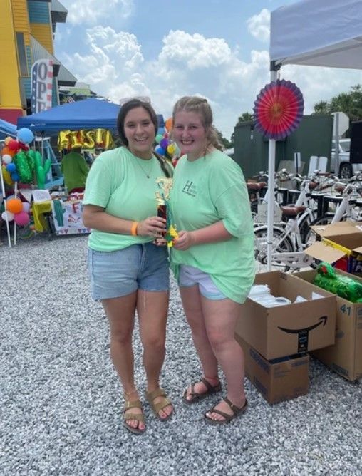 Two women in green shirts are standing next to each other in a gravel area.