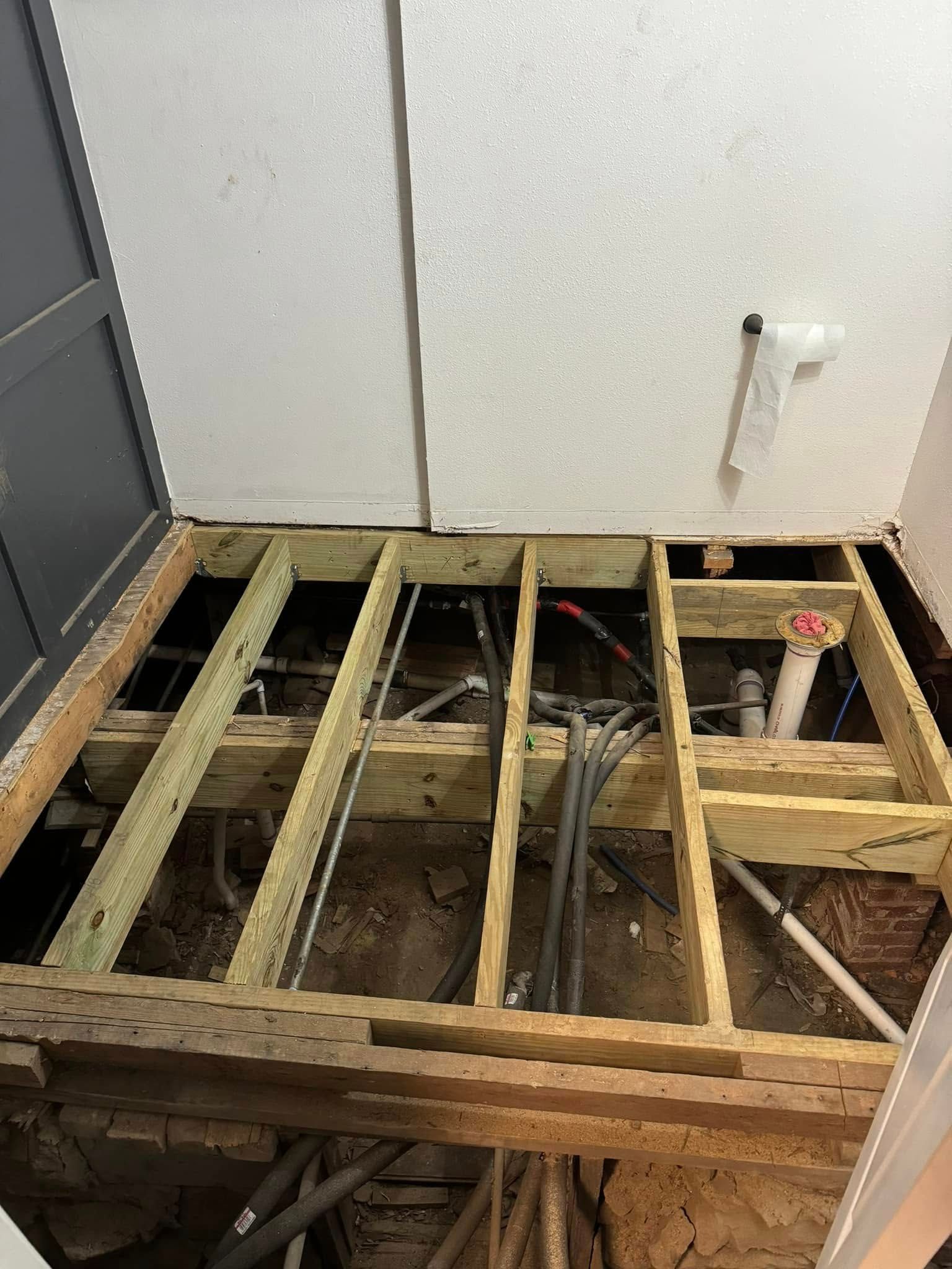 Wooden floor frame exposed, showing pipes and electrical wiring, likely a bathroom renovation in progress.