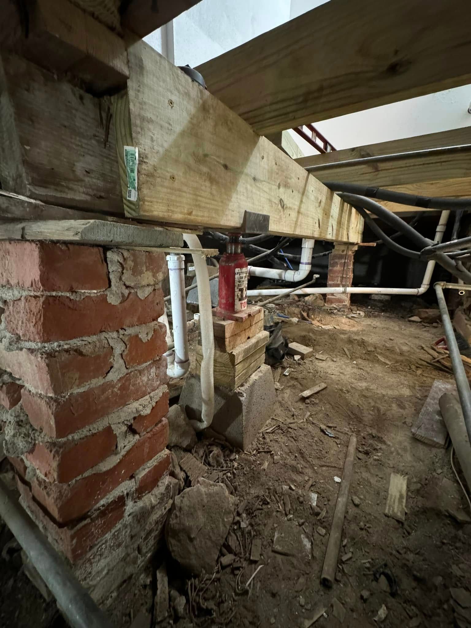 Foundation repair: brick supports a wooden beam, with plumbing and wiring visible underneath a house.