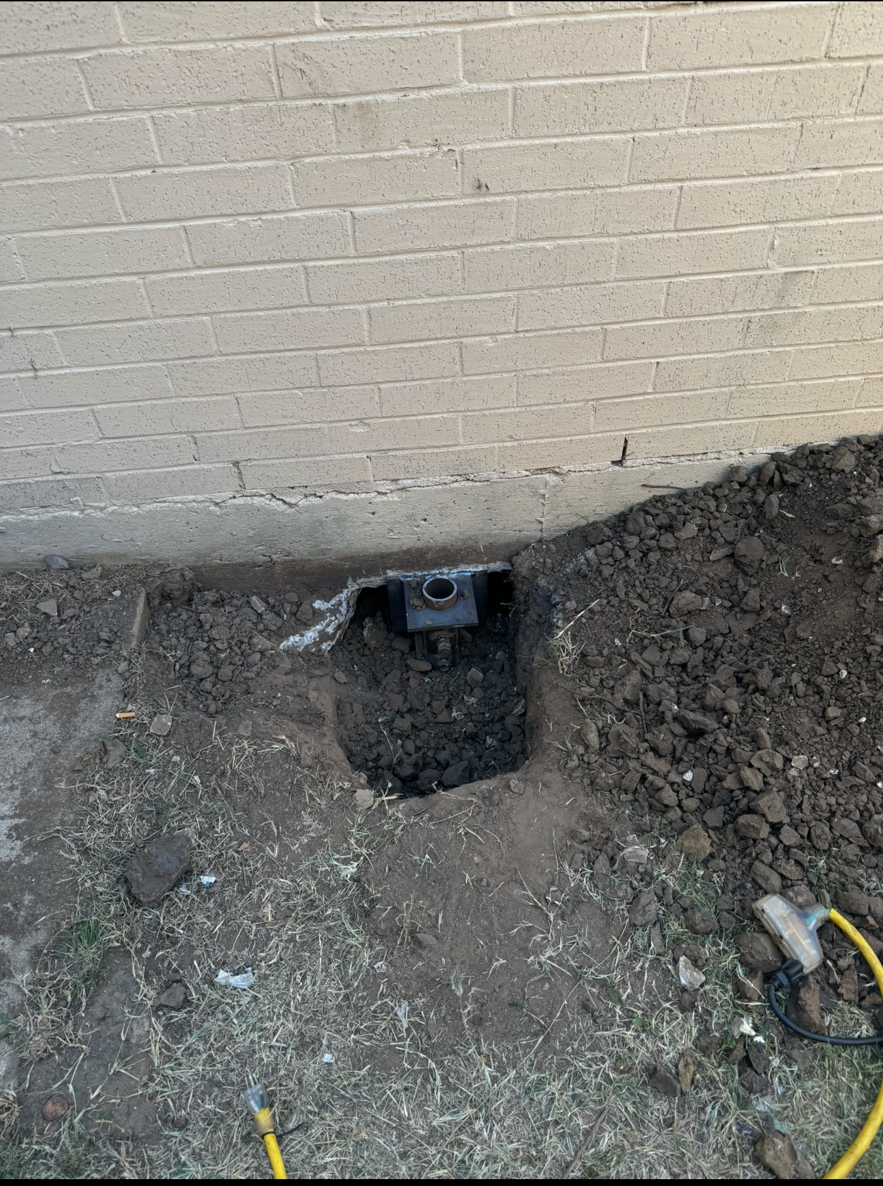 Hole dug near a cinder block wall, revealing a black pipe or fitting. Earth and grass surround the hole.