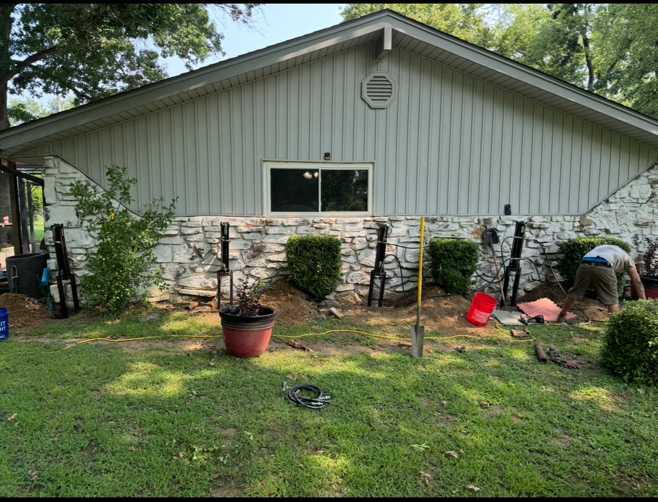 Exterior of a home; workers are digging in the yard. Stone wall and white siding. Green grass and shrubbery.