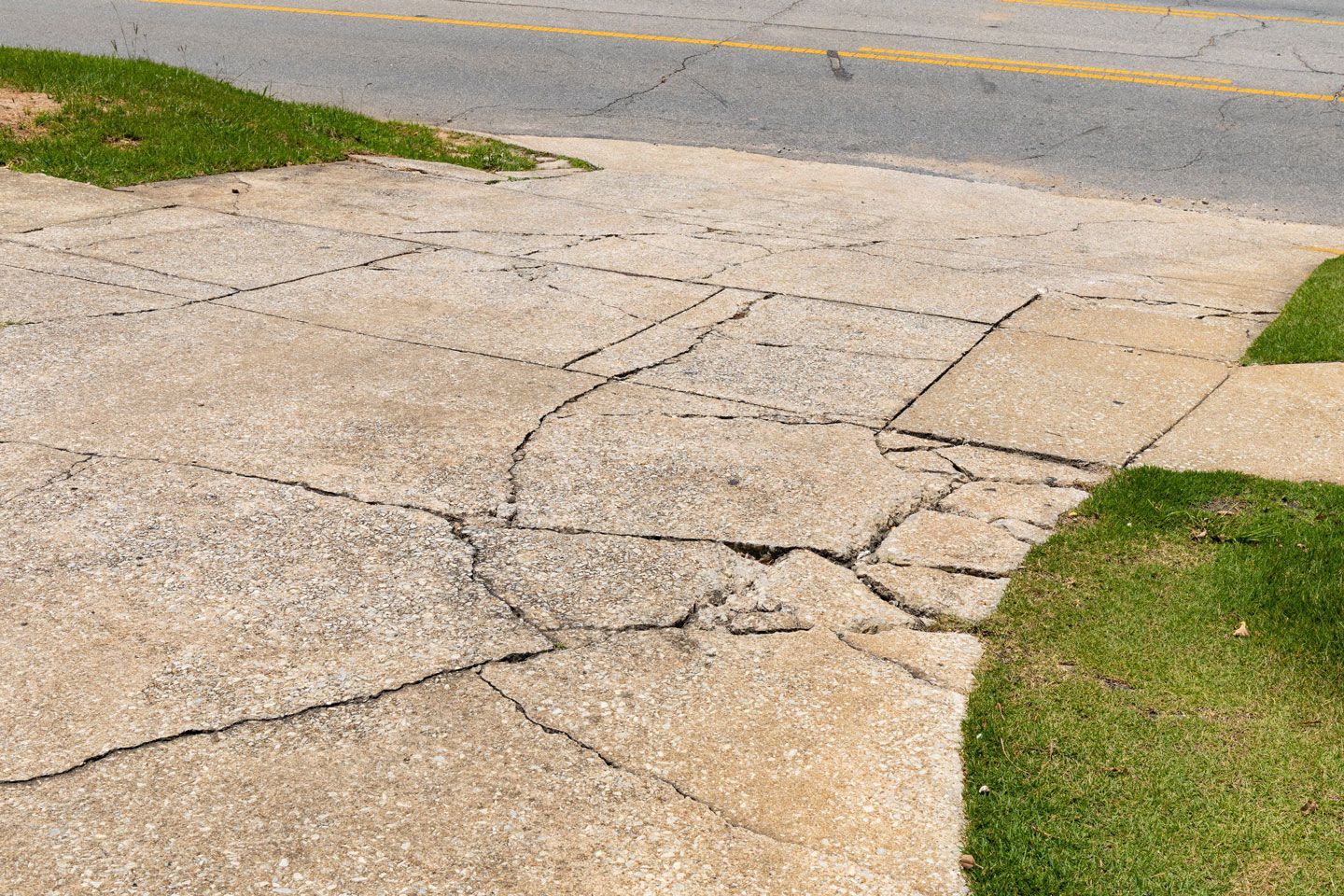 Cracked and broken concrete sidewalk next to a road and grassy area.