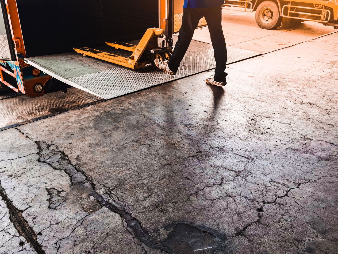 Person walking off truck bed toward cracked concrete. Yellow forklift, orange truck, and gray pavement.