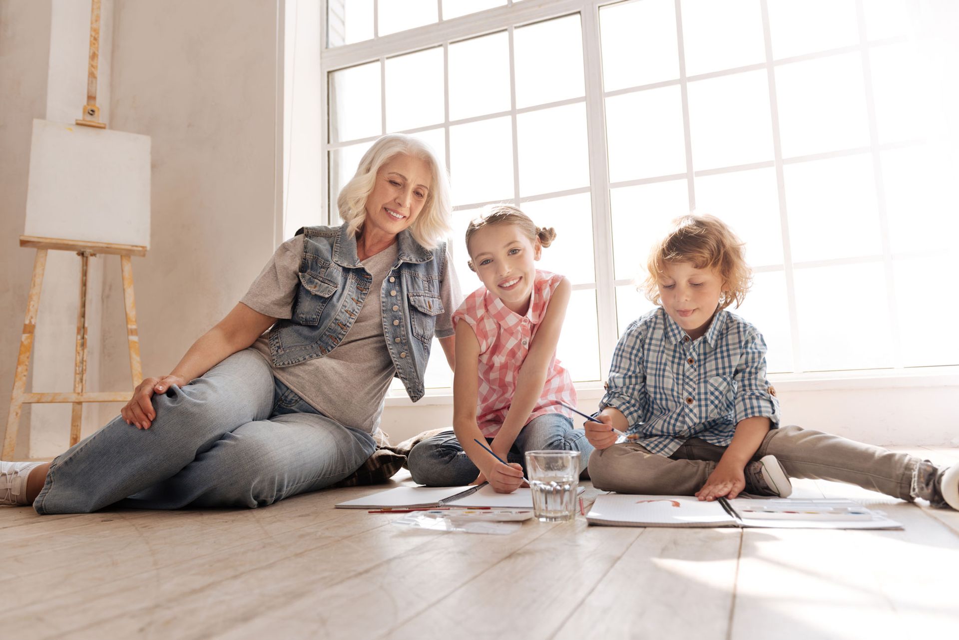 Woman and two children smiling while sitting on floor painting in front of a window.