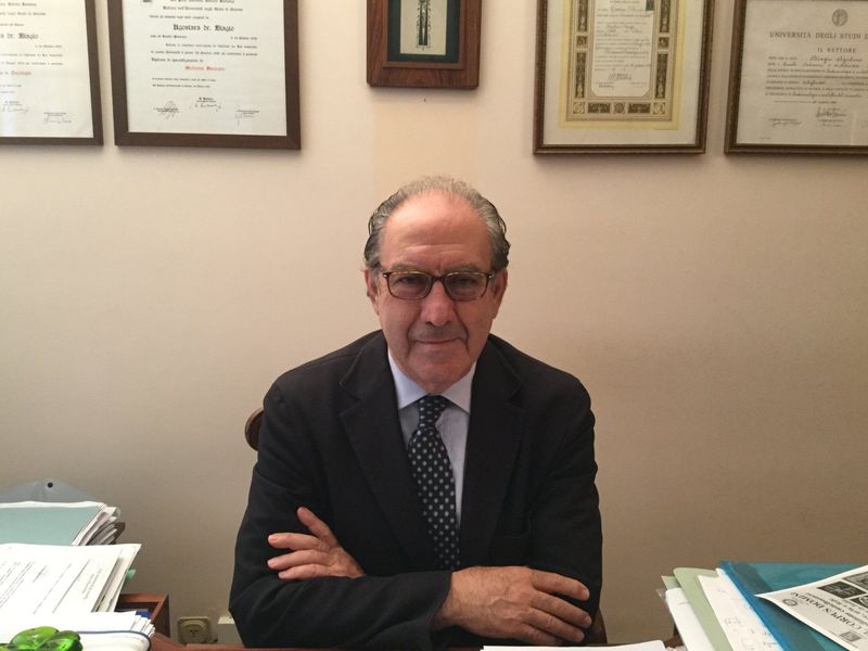 Uomo in giacca e occhiali, seduto alla scrivania, braccia incrociate, documenti incorniciati sullo sfondo.