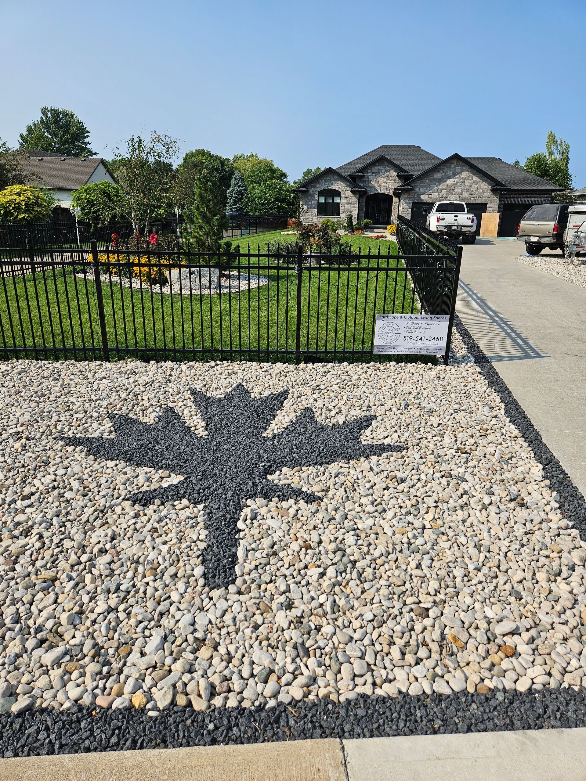 Gravel landscaping with a dark rock maple leaf design in front of a house with black fencing.