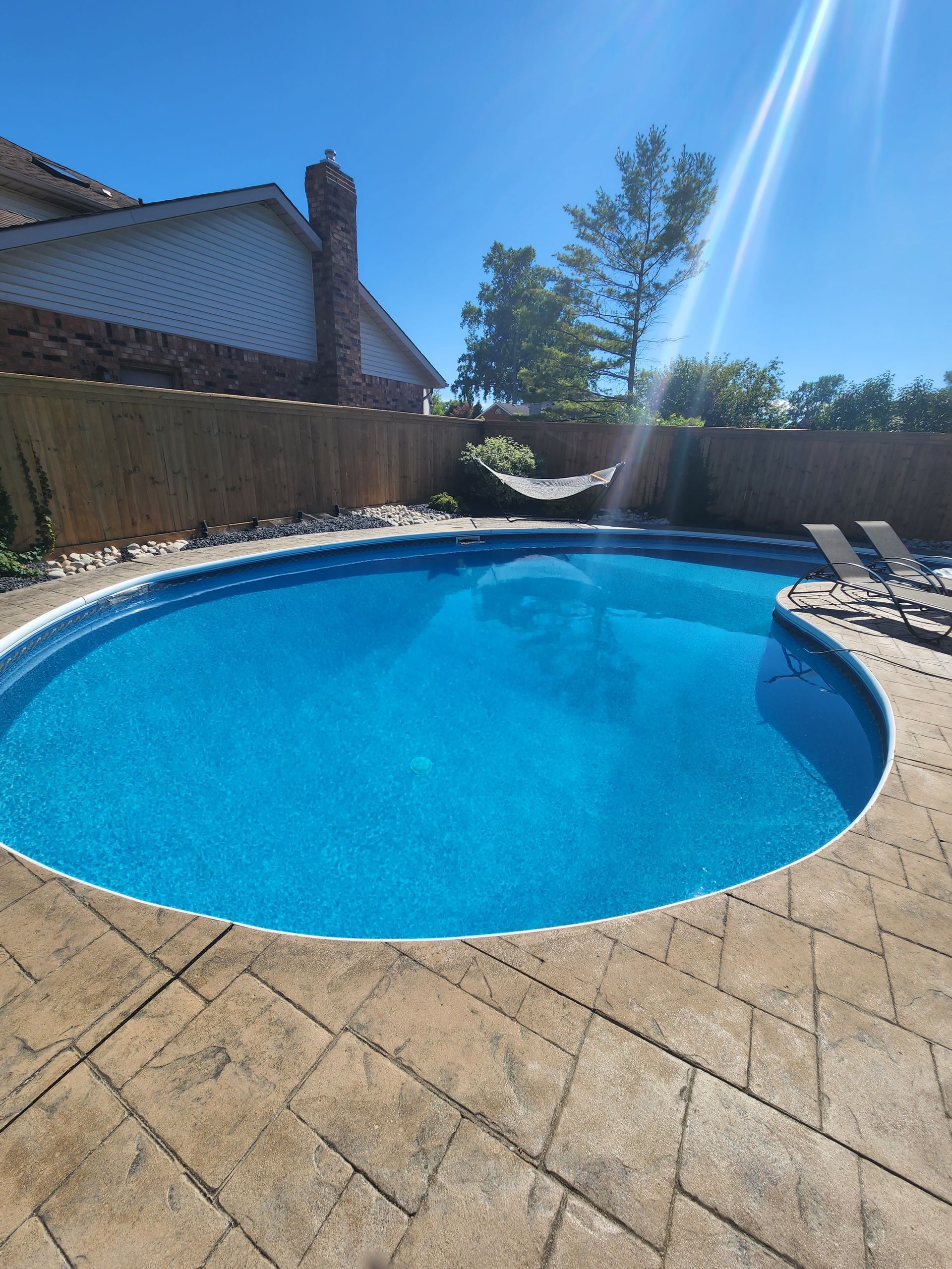 A sunlit backyard with an oval-shaped swimming pool, patterned concrete decking, and a wood fence under a clear blue sky.