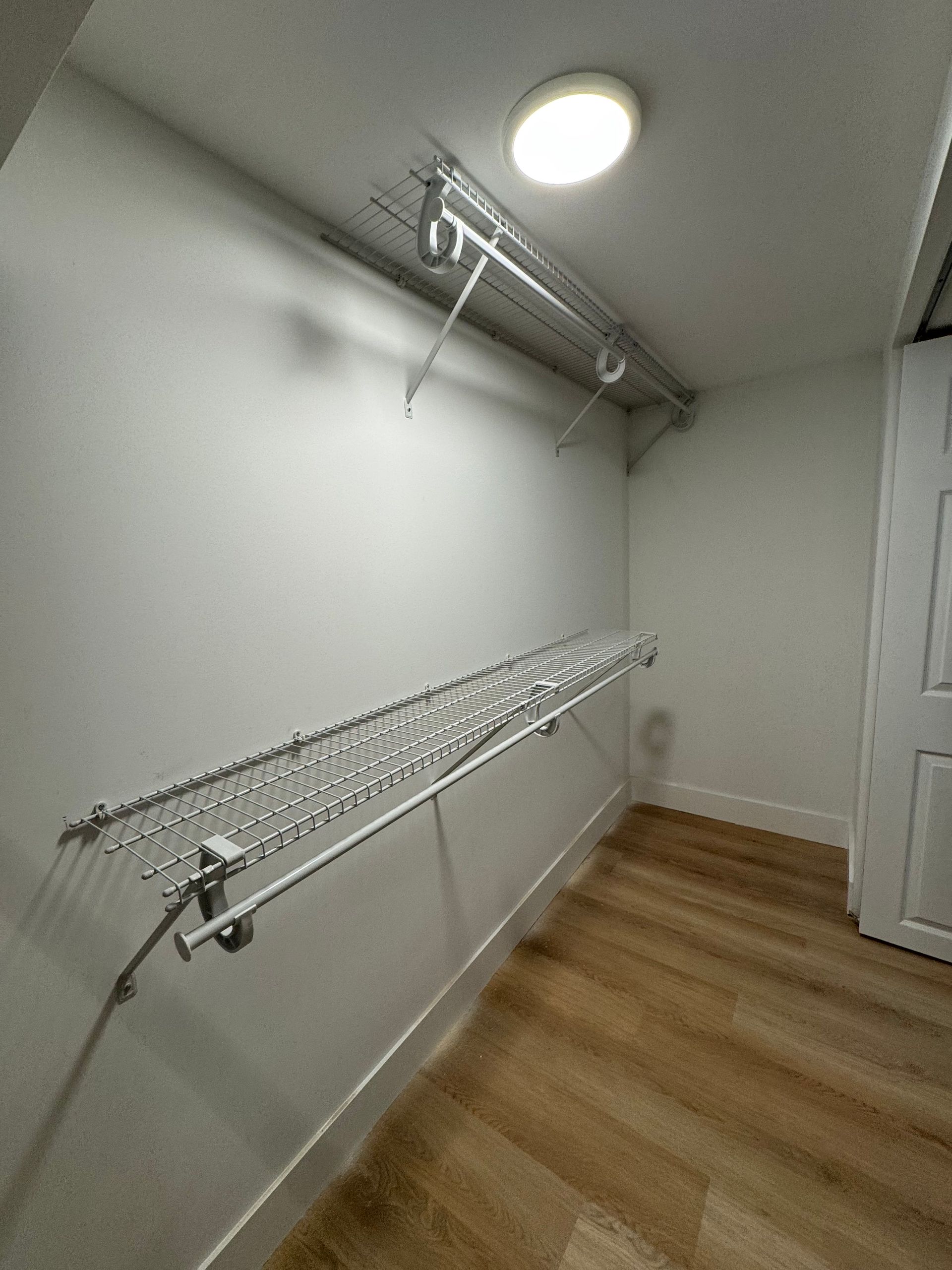 Empty closet with white walls, wood floor, ceiling light, and wire shelves on both sides.