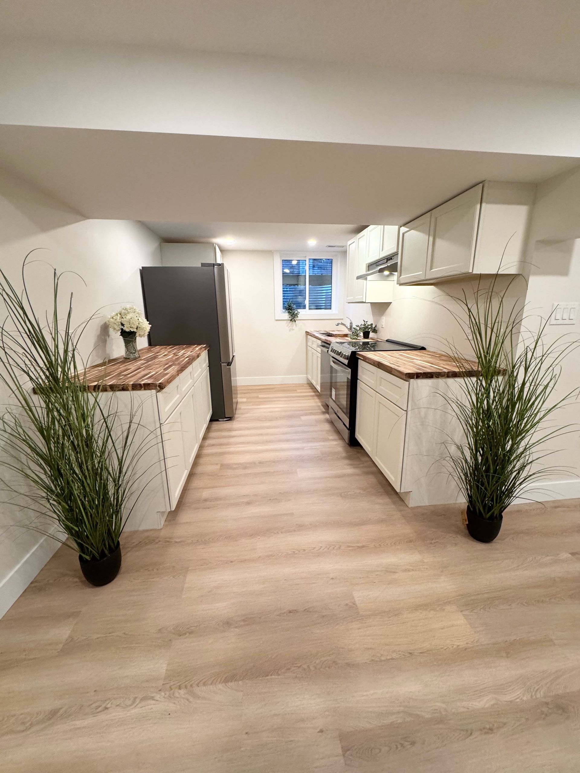 Bright narrow kitchen with white cabinets, wood countertops, light flooring, and tall potted plants at the entrance