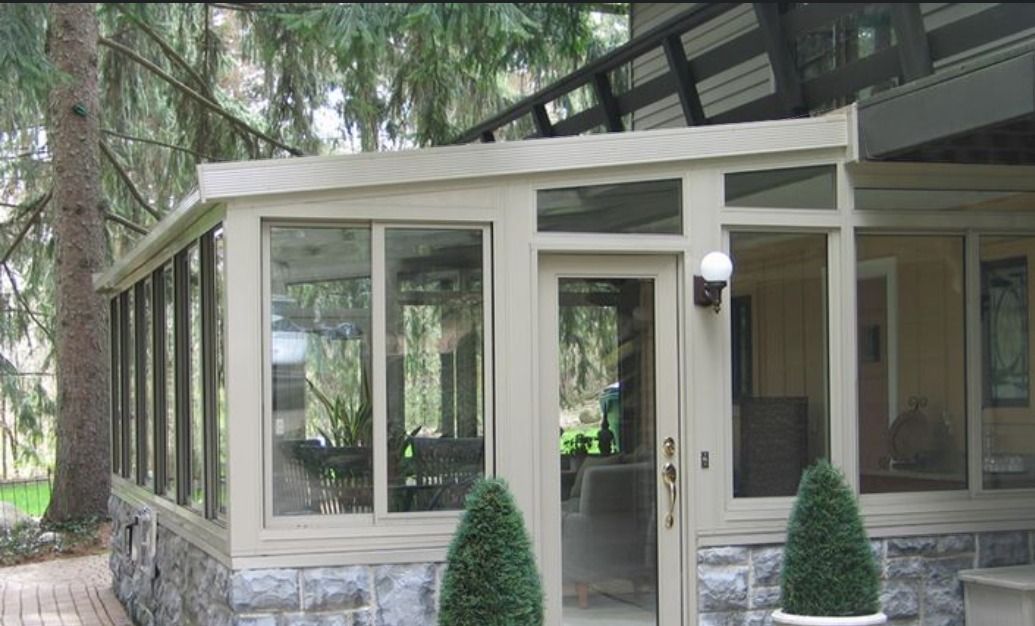 A sunroom with large glass panels and light-colored frames, set against a stone base with two small evergreen shrubs.