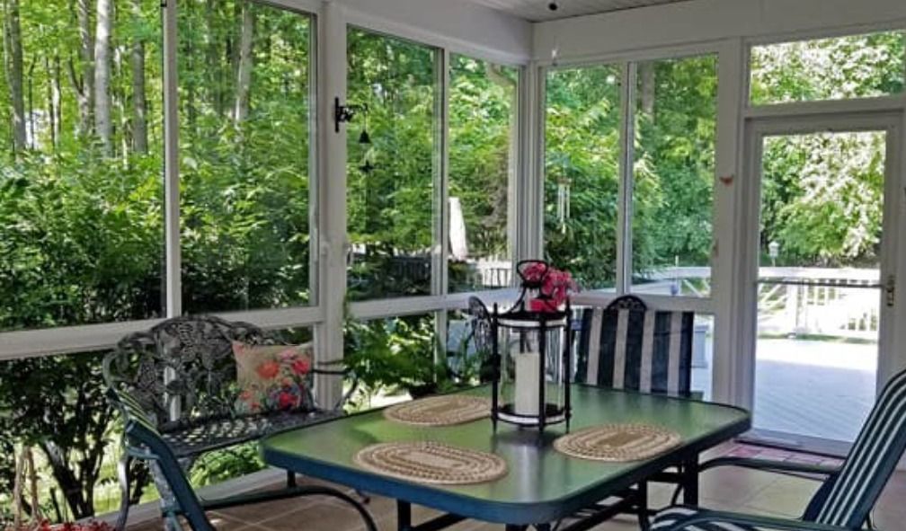 A sunroom with a dining table, chairs, and a metal bench, overlooking a lush green forest through large glass panels.