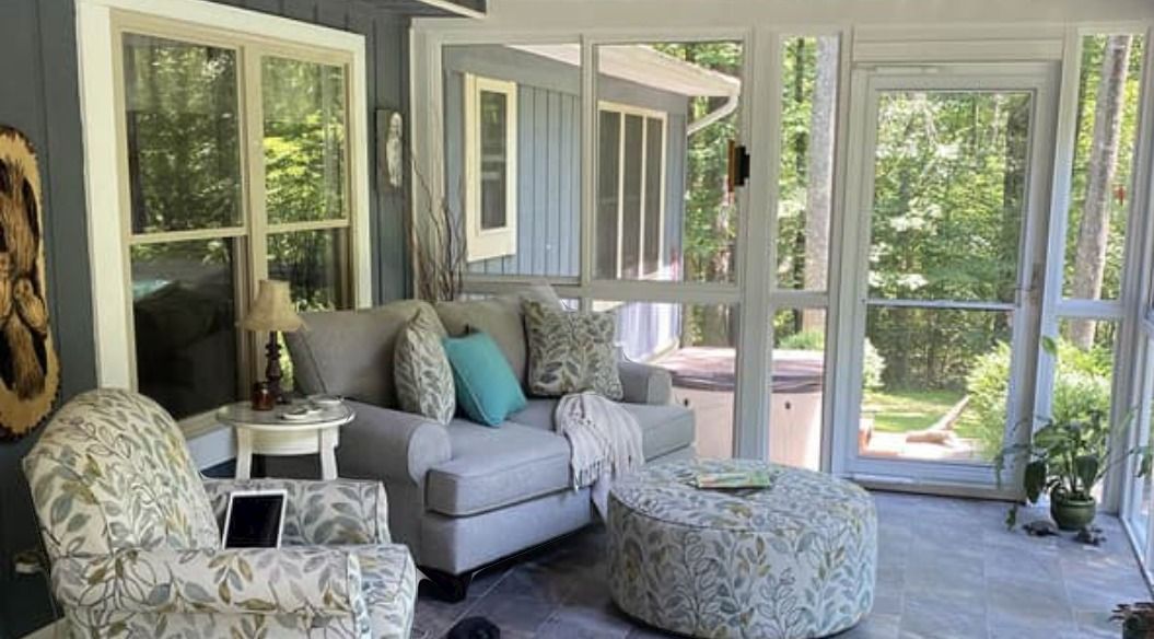 A cozy screened-in sunroom with a grey sofa, matching patterned armchair, and round ottoman, overlooking green trees.