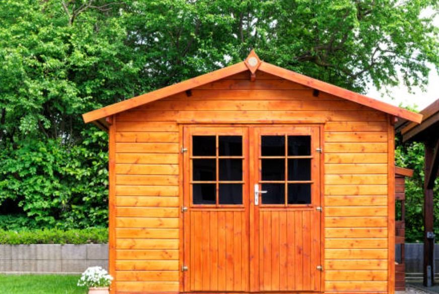 A light-colored wooden garden shed with double doors and a gabled roof, situated outdoors in front of a leafy green hedge.