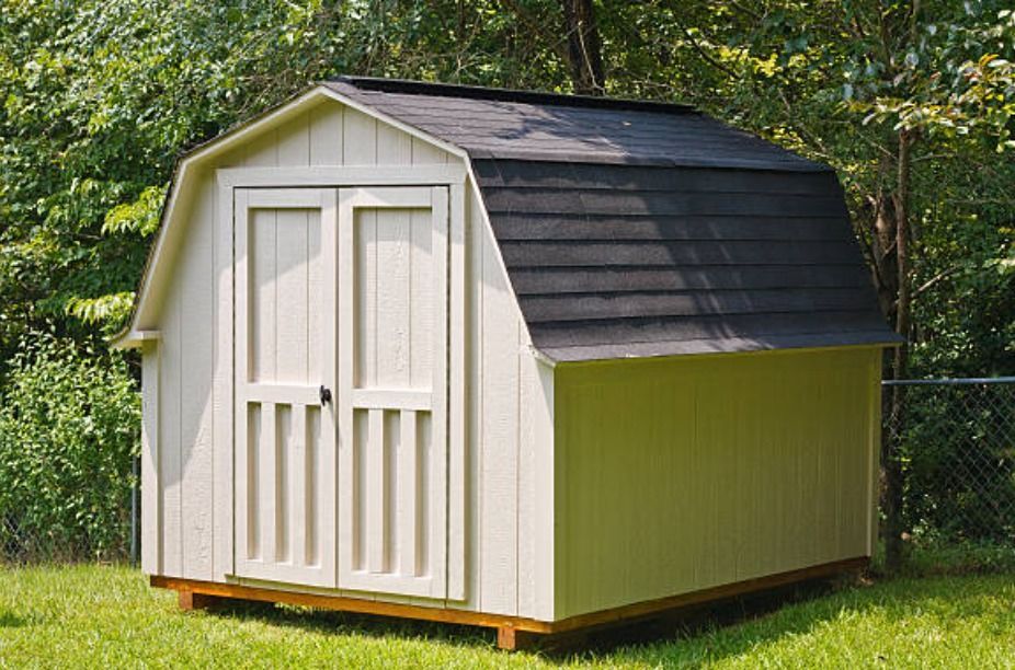 A small, cream-colored wooden storage shed with a black gambrel roof, set on a grass lawn in front of trees.
