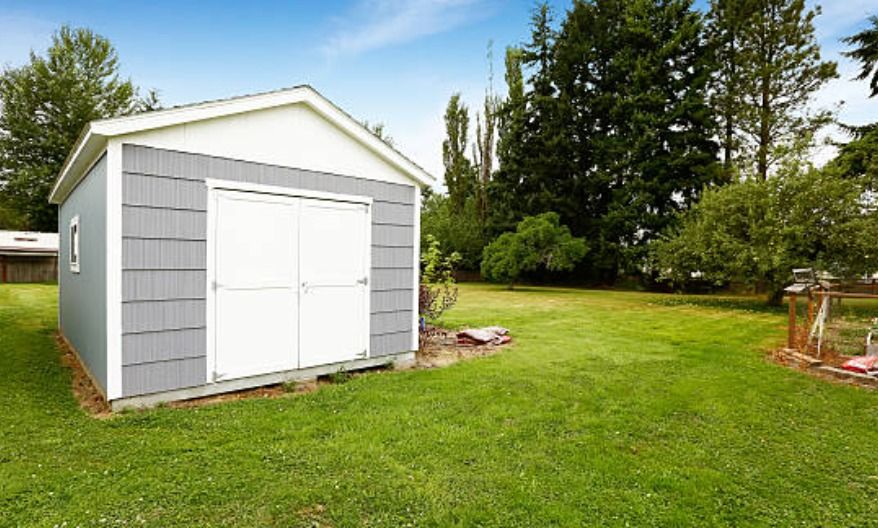 A grey shed with white trim and double doors sits on a green lawn in a yard with trees and a small wooden structure.