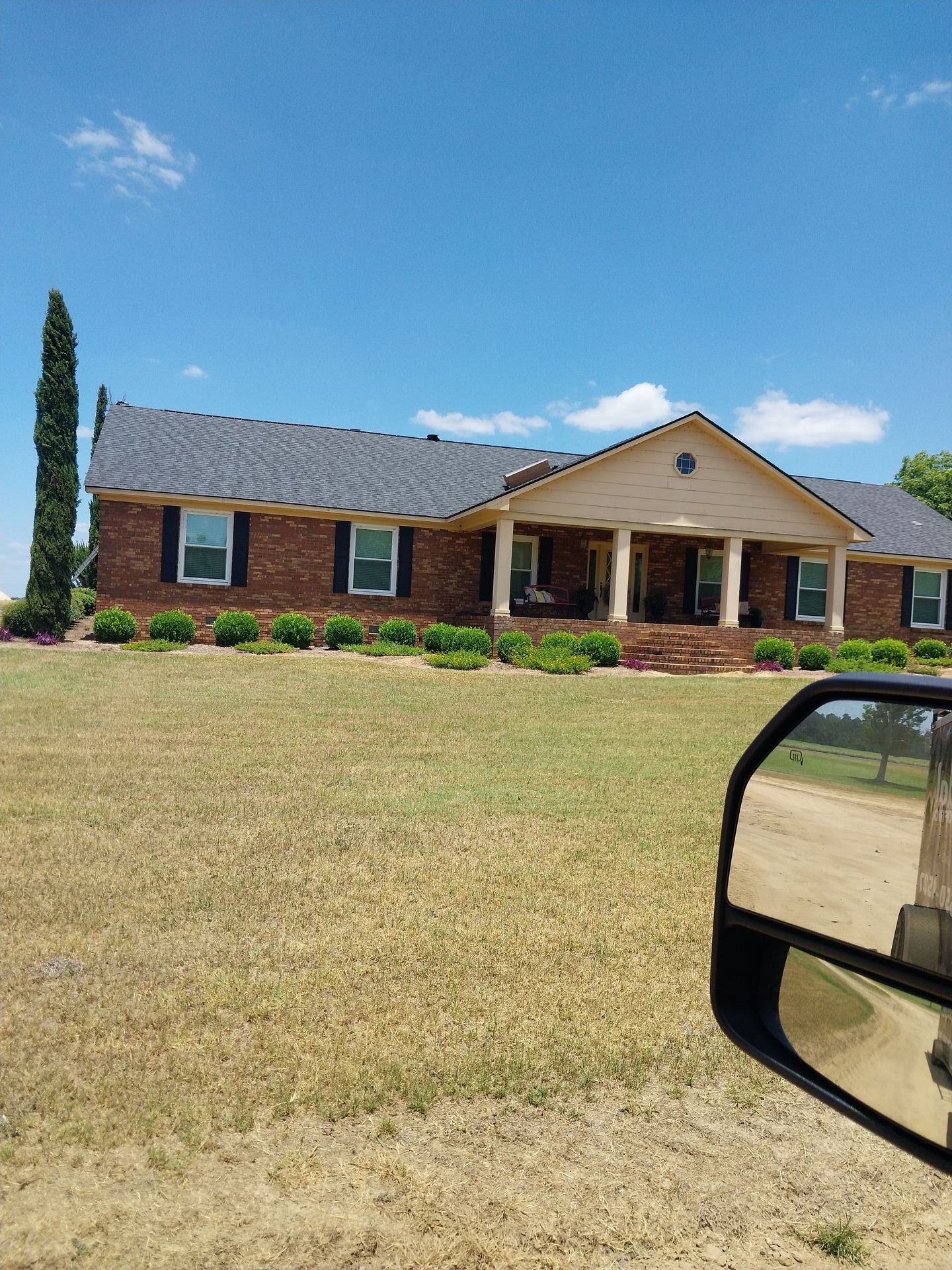 Brick ranch house with black shutters and columns, set on a large lawn under a blue sky.