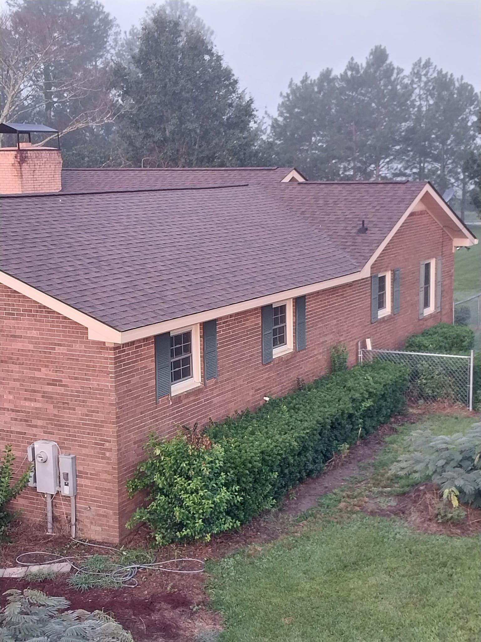 Brick house with brown roof, green shutters, and a hedge row in a yard; misty background.