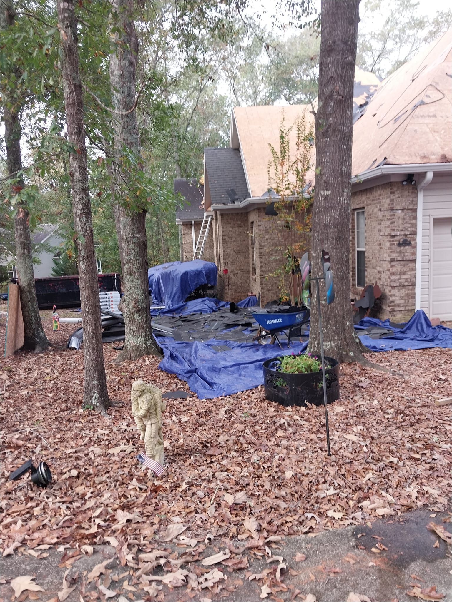 House with roof being worked on, covered with blue tarps. Yard scattered with leaves, trees.