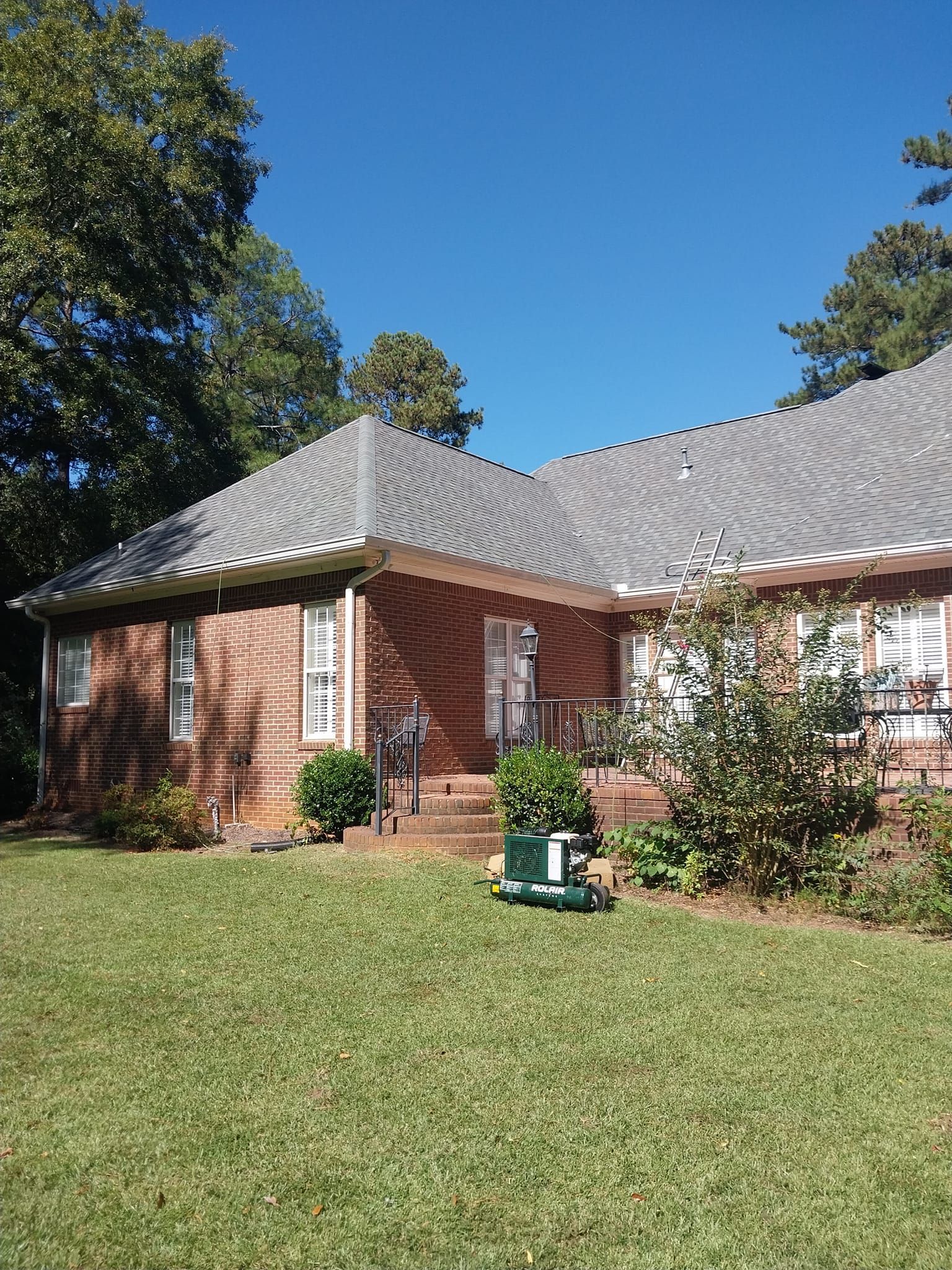 Brick house with gray roof, green grass, and trees under a clear blue sky.