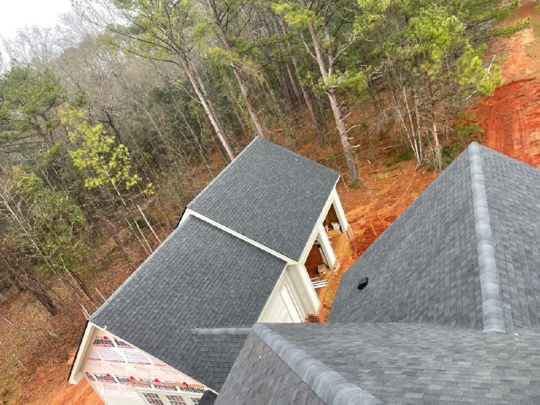 View of a house with a dark gray shingled roof, set against a forest and a red-earth hillside.