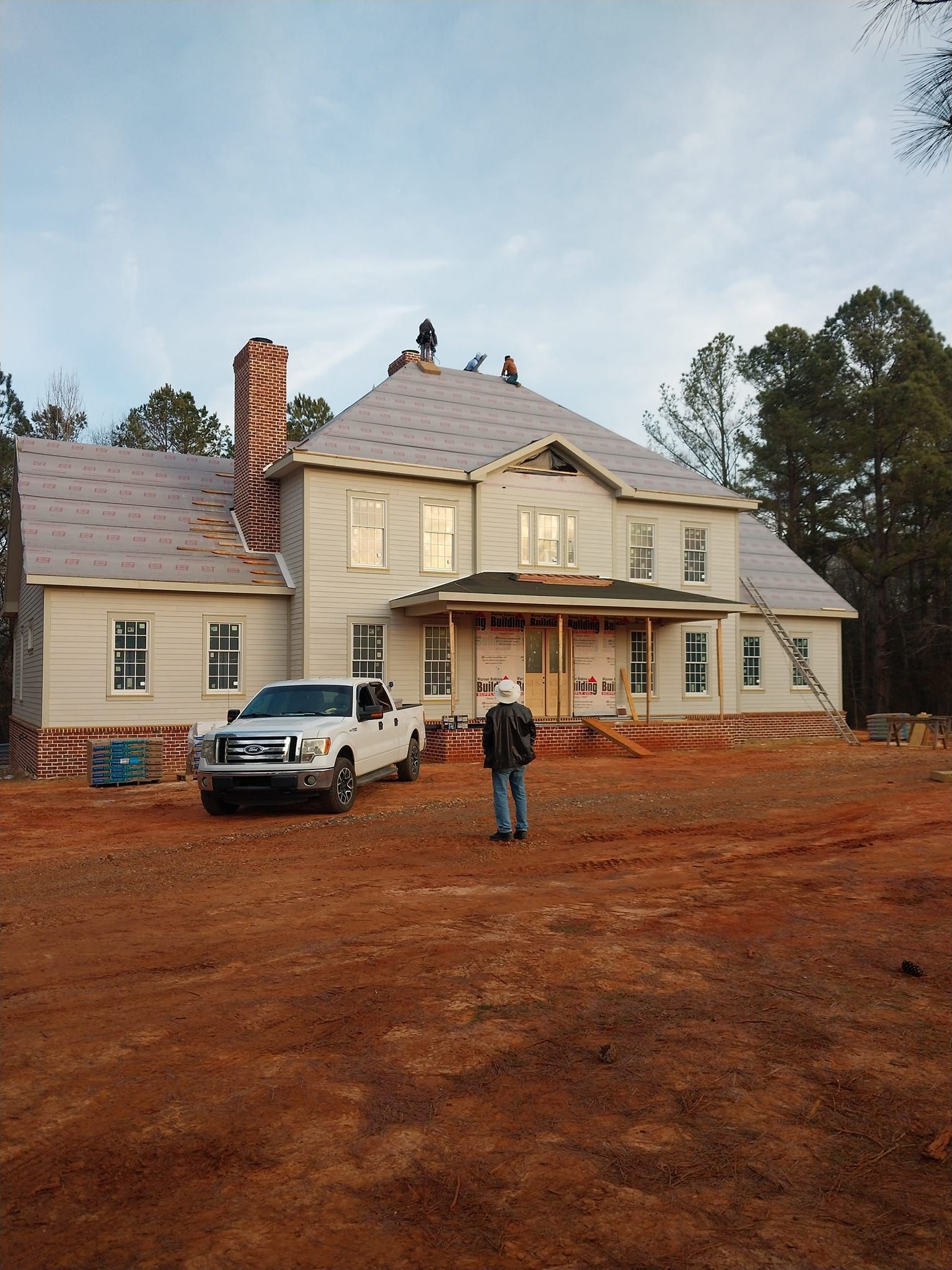 House under construction, workers on roof, truck in front. Red clay soil, overcast sky.