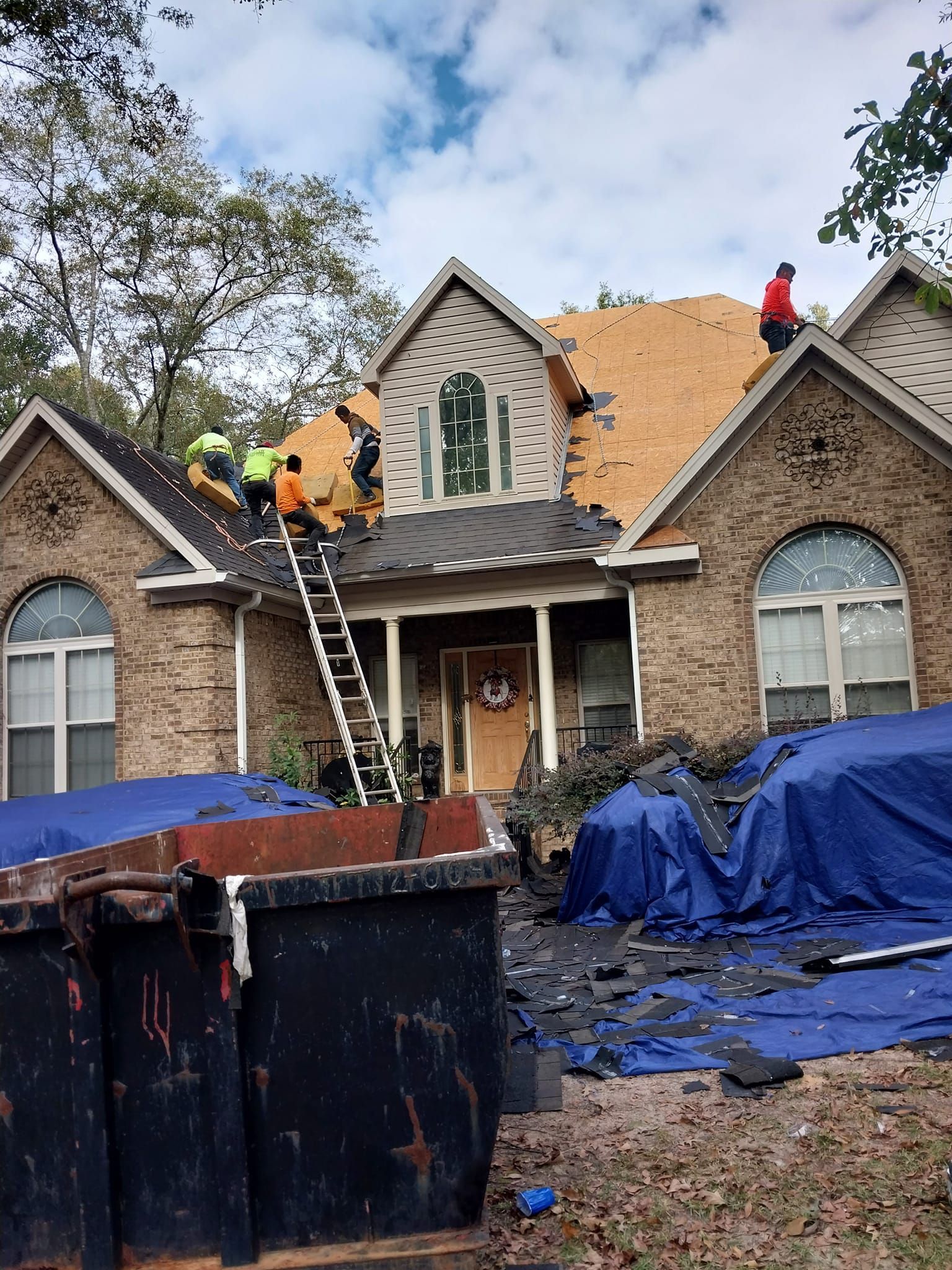 Roofers replacing shingles on a brick house. Debris and dumpster in the foreground; workers on the roof.