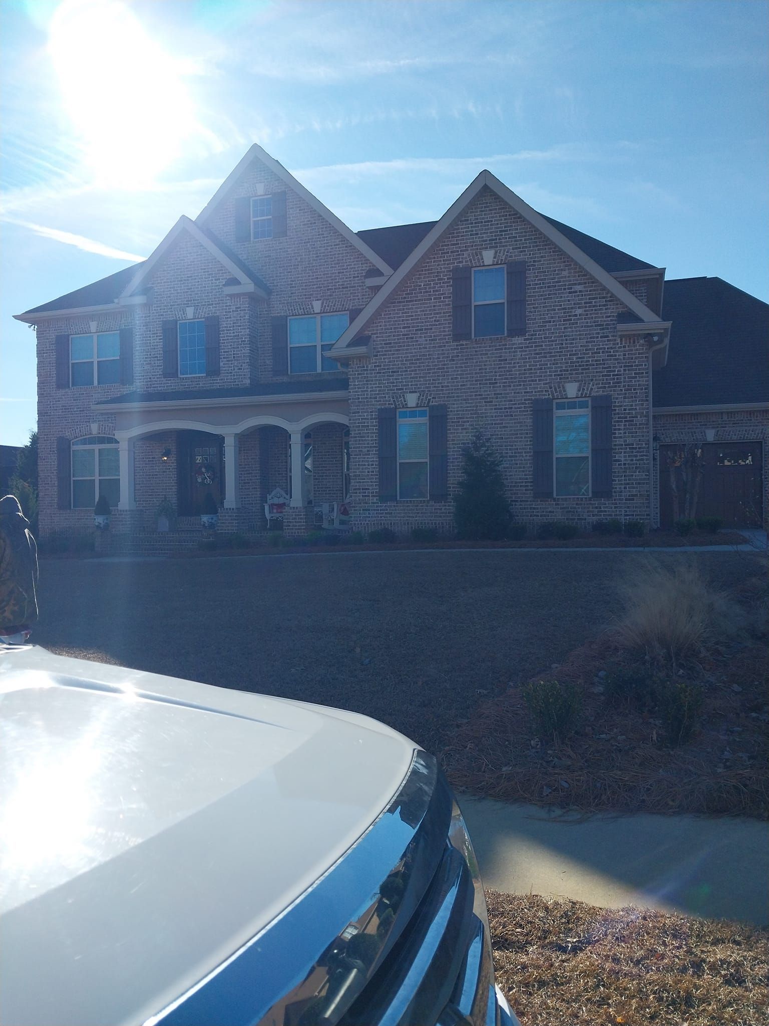 Two-story brick house with dark shutters, under a bright sun. A silver vehicle is in the foreground.