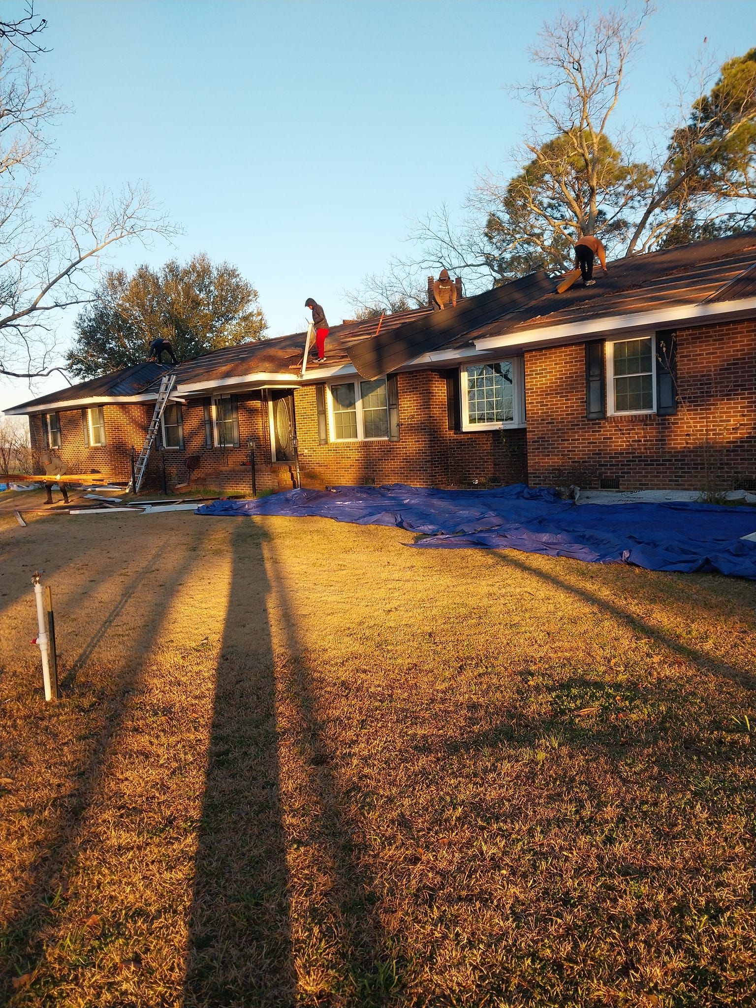 House with tarp-covered roof; person on roof, sun casting long shadow on grass.