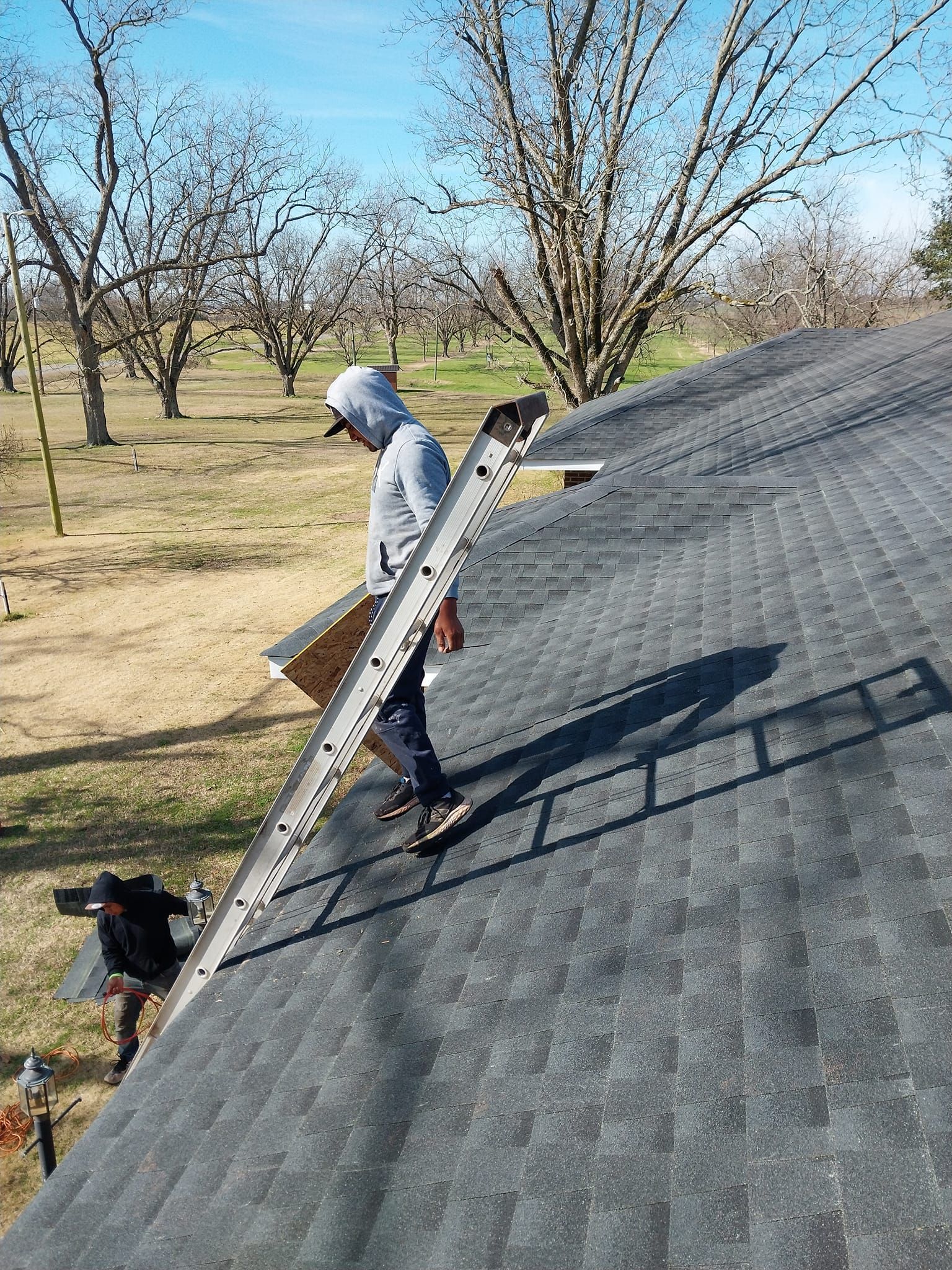 Two people on a roof with a ladder. One person carries a piece of wood. Outdoors, sunny day.