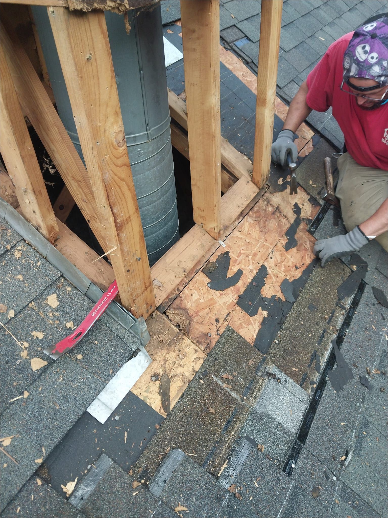 Roofer repairs roof around a chimney. They're removing old shingles and working on a wooden frame.