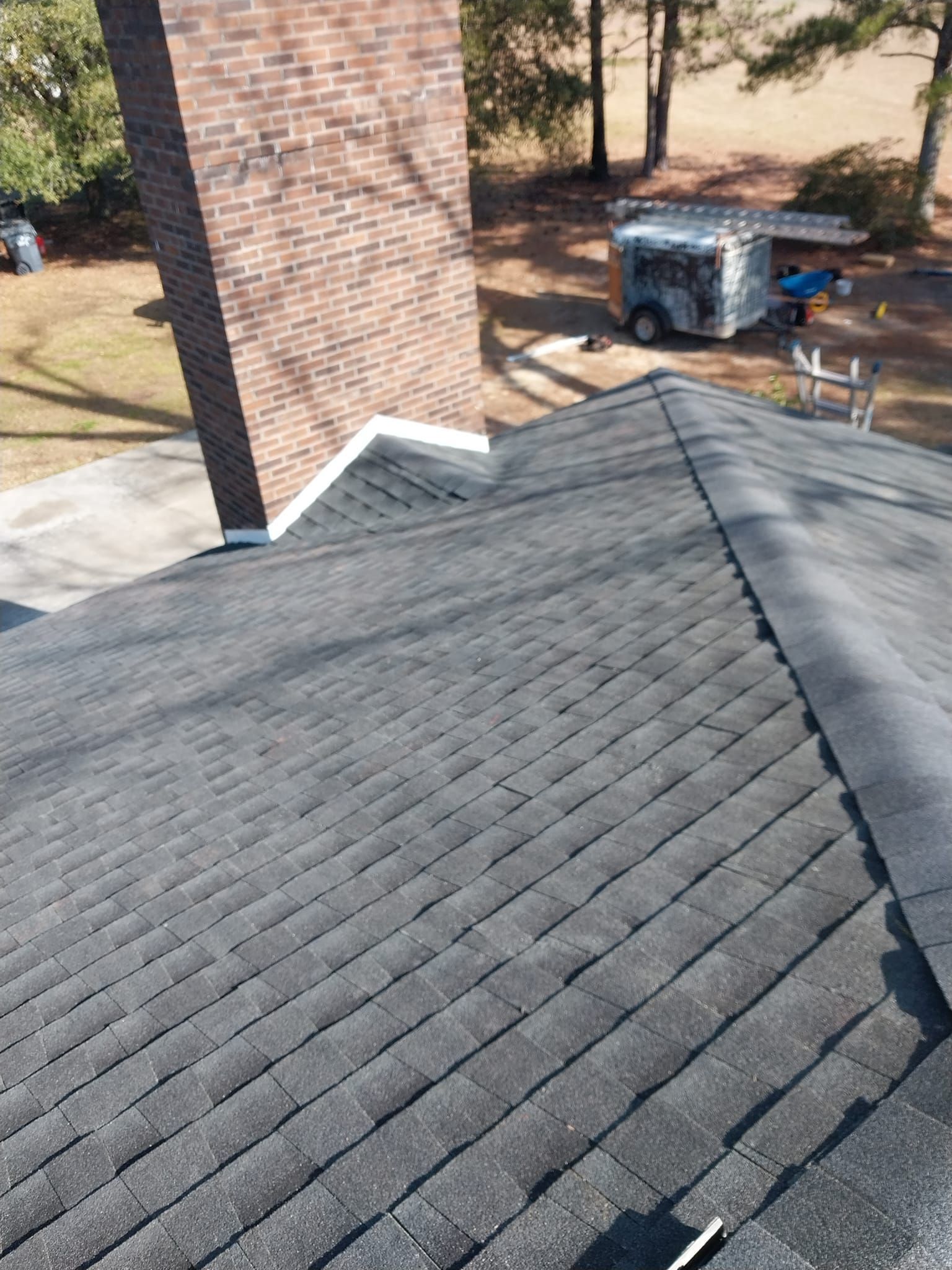Roof with dark gray shingles, a brick chimney, and a trailer in a grassy yard.