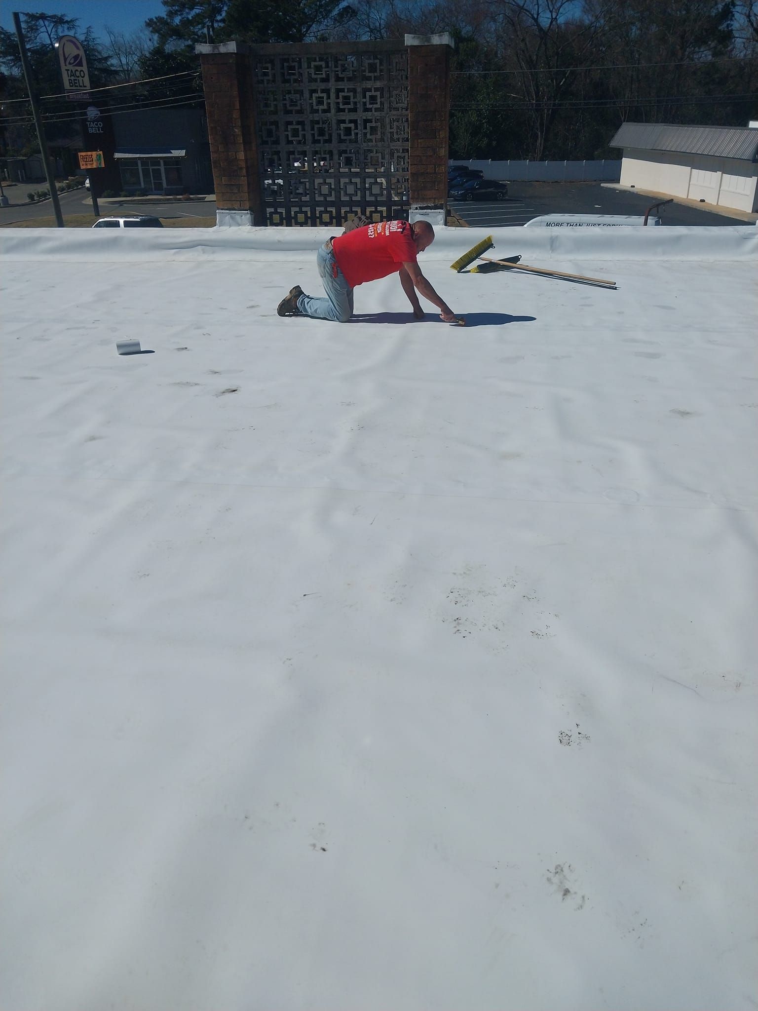 Man on a white roof, kneeling, working with tools. Brick building in the background on a sunny day.