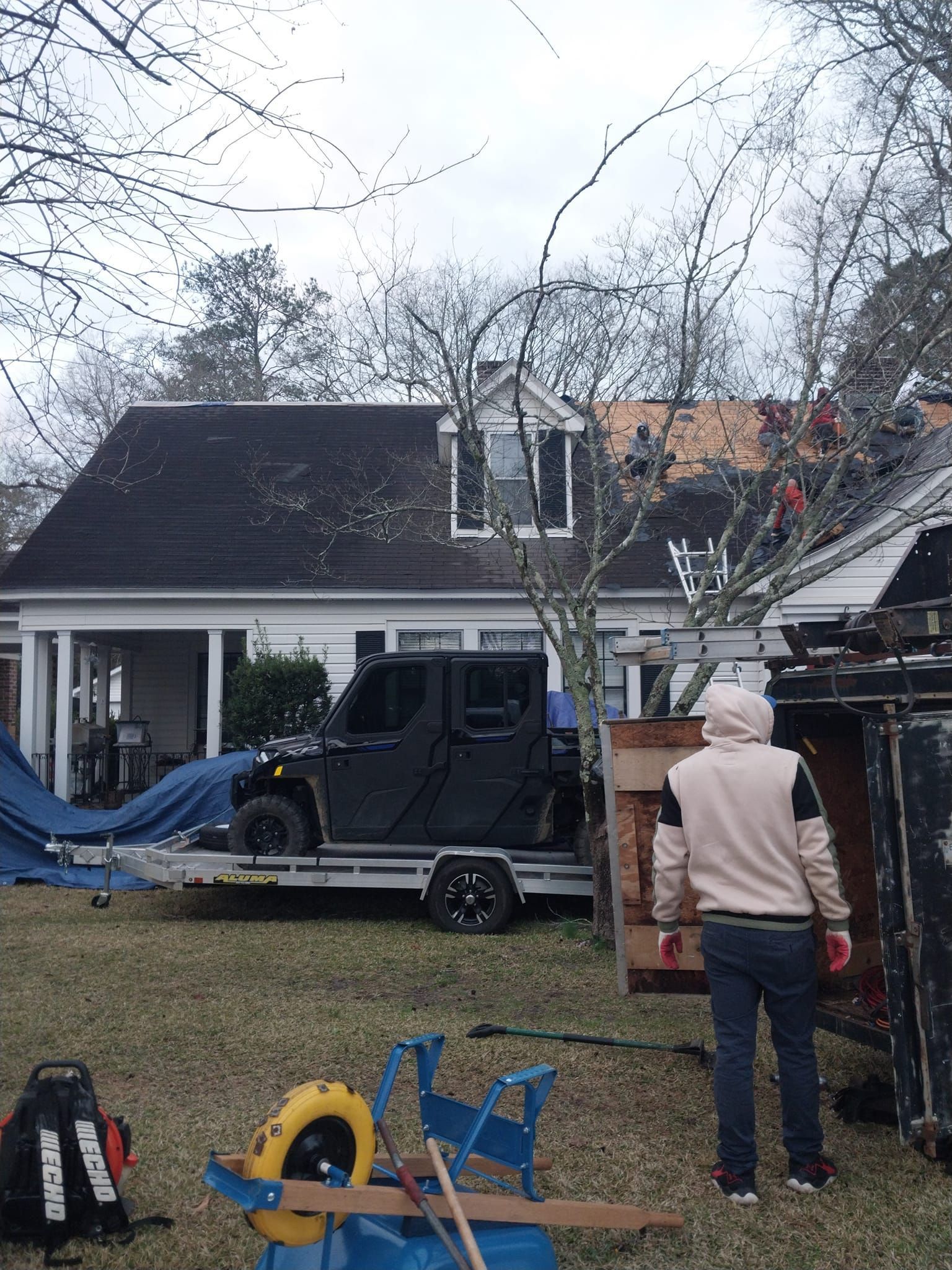 Person near a dumpster; UTV on a trailer; house in the background, tarp on roof.