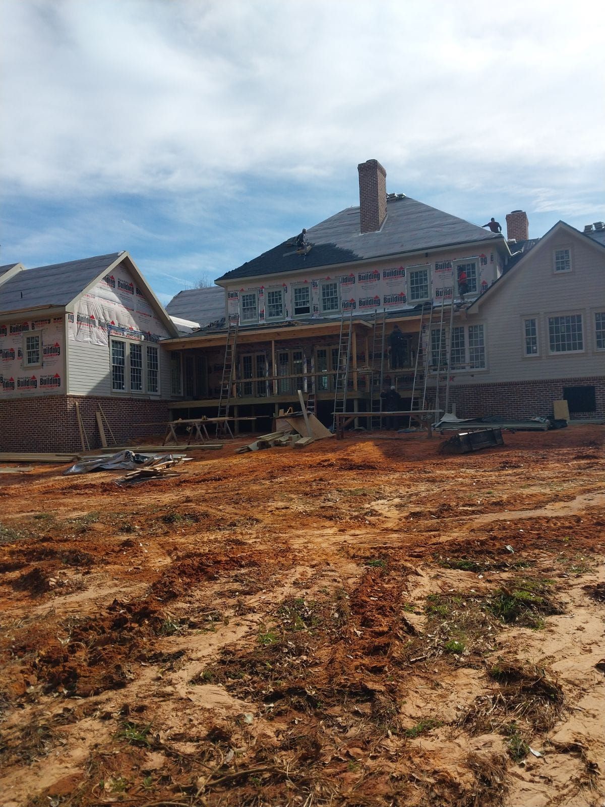 House under construction with exposed framing and wrap, set in a dirt lot.