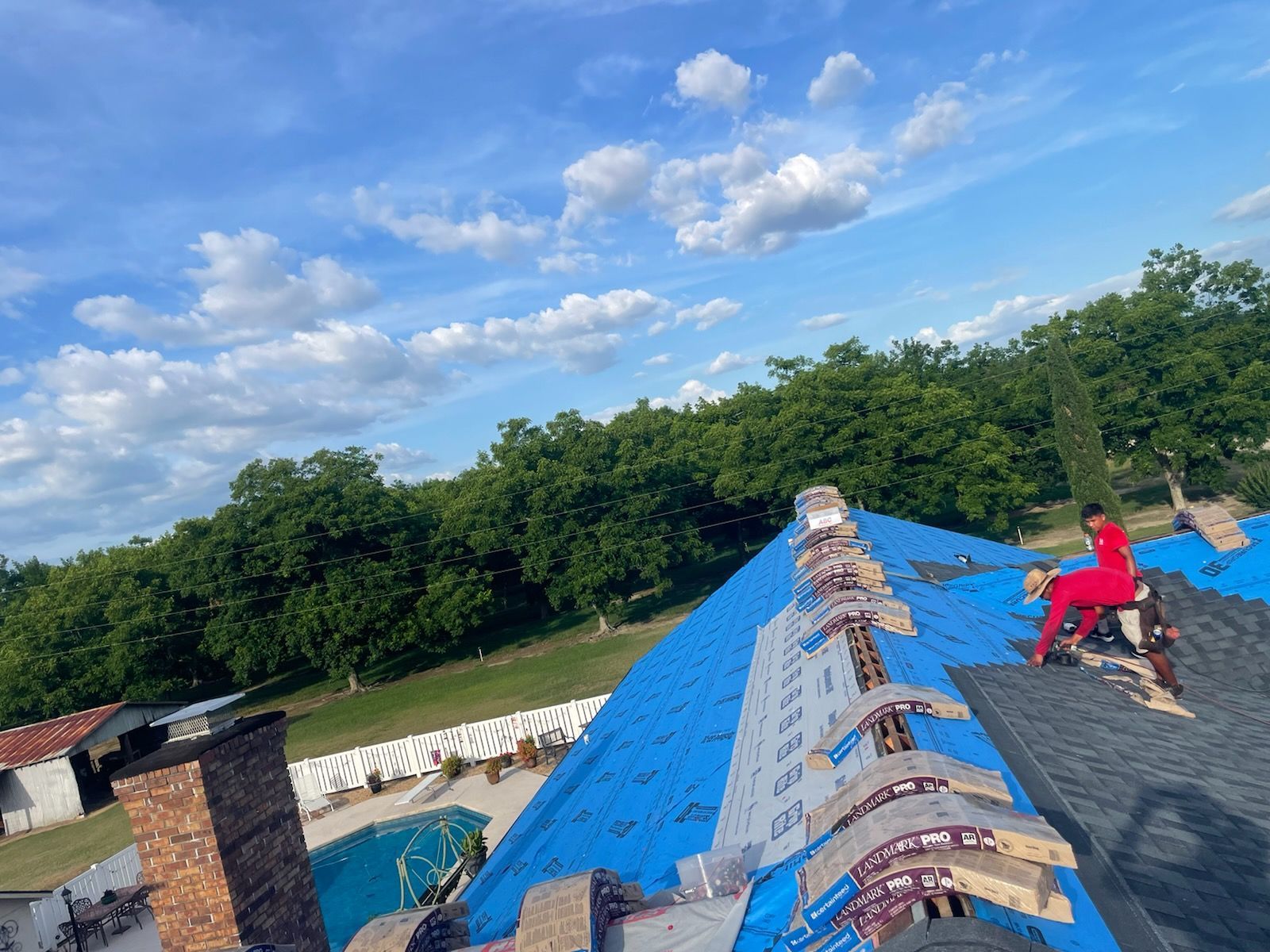 Roofer on a partially shingled roof under a blue sky.  Trees in the background.