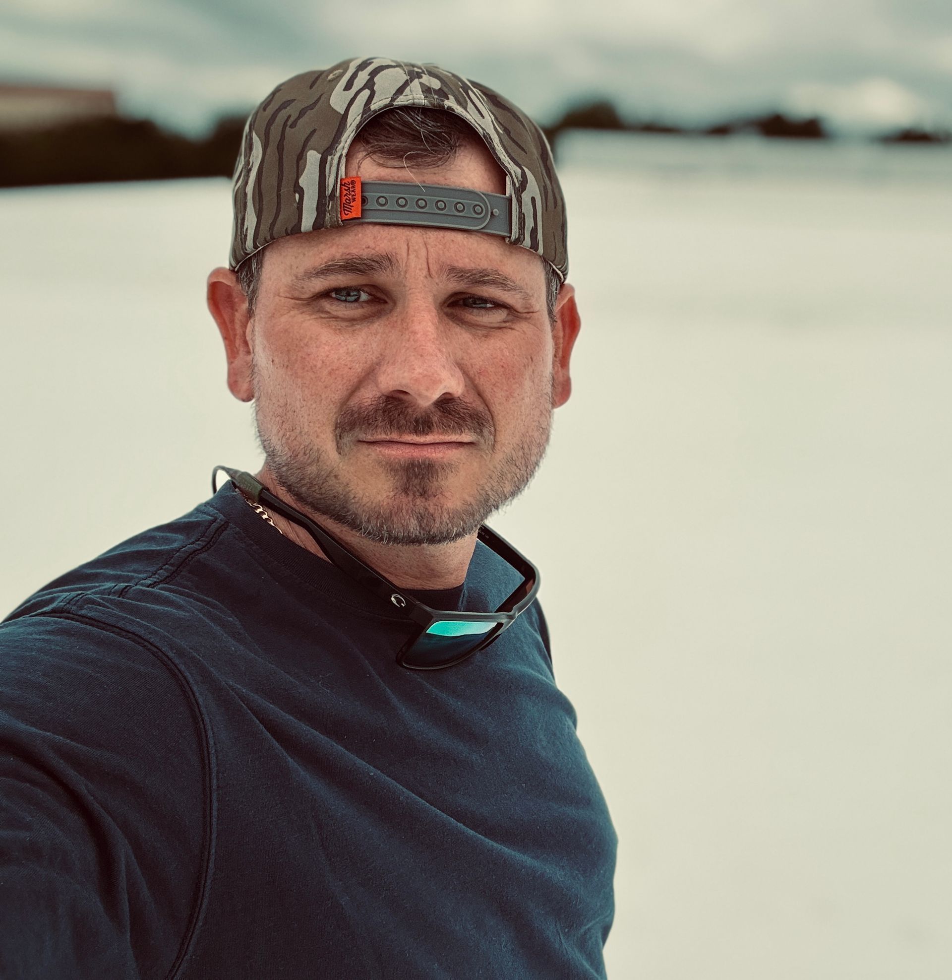 Man in camo hat and blue shirt, looking at the camera outdoors, sunglasses around his neck.