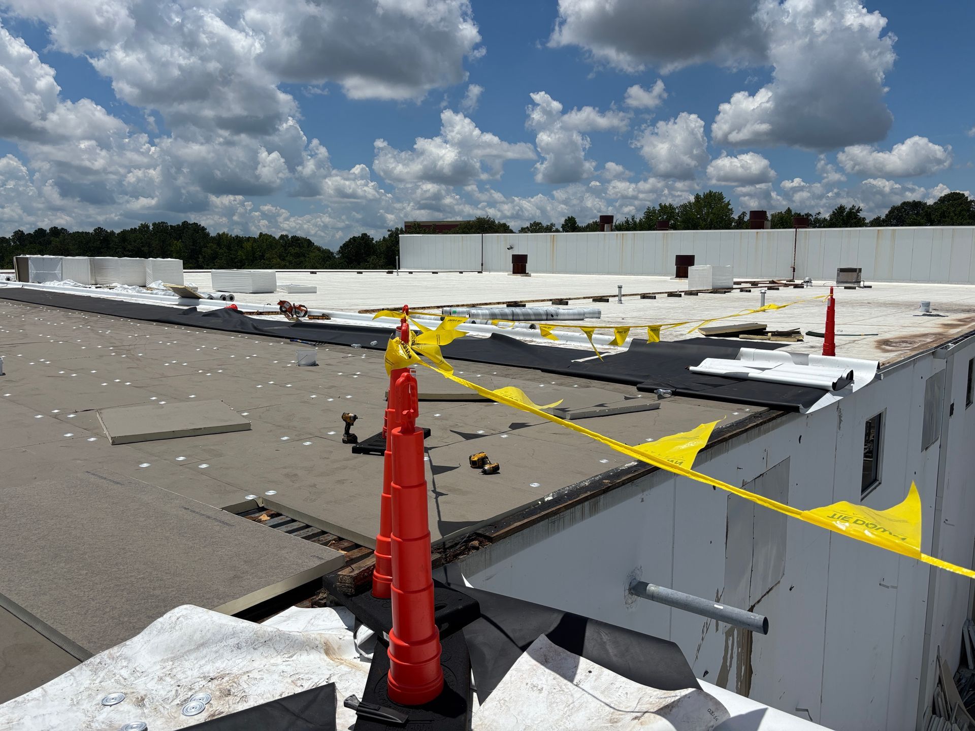 Rooftop with torn roofing material, safety cones, and caution tape under a partly cloudy sky.