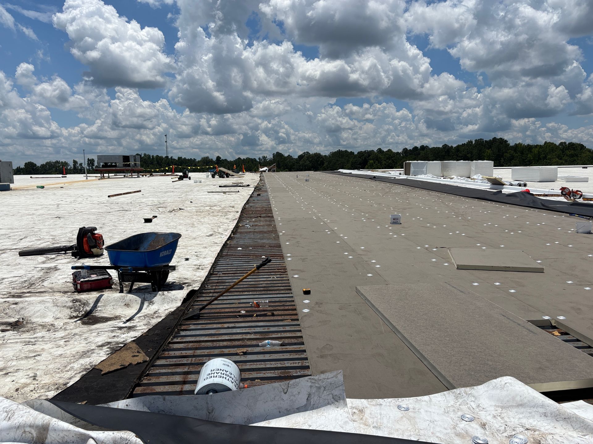 Roofing work in progress on a large flat roof, showing exposed insulation and materials under a partly cloudy sky.
