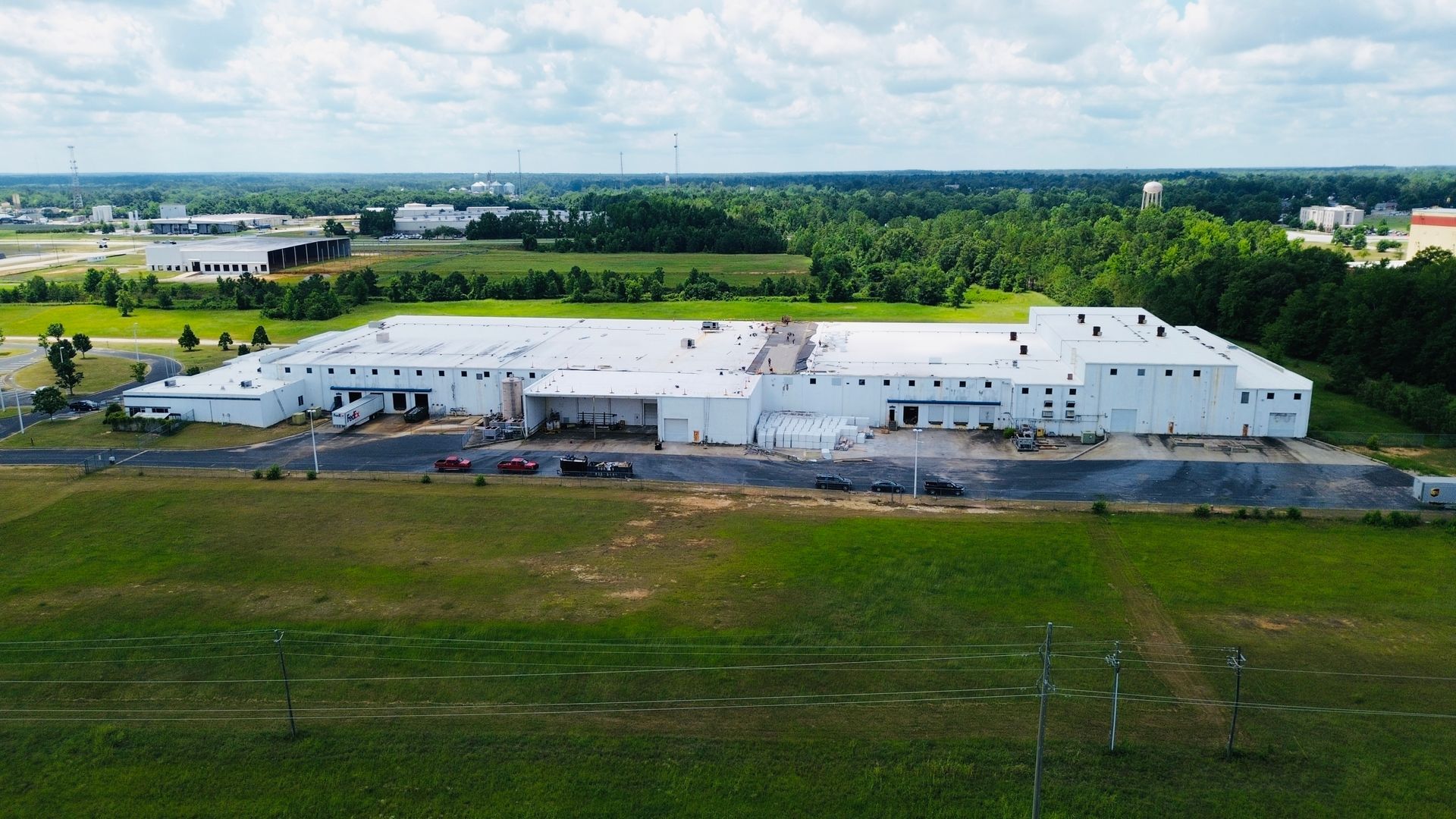 Large white industrial building with flat roof and loading docks, set in a green field with trees in the background.