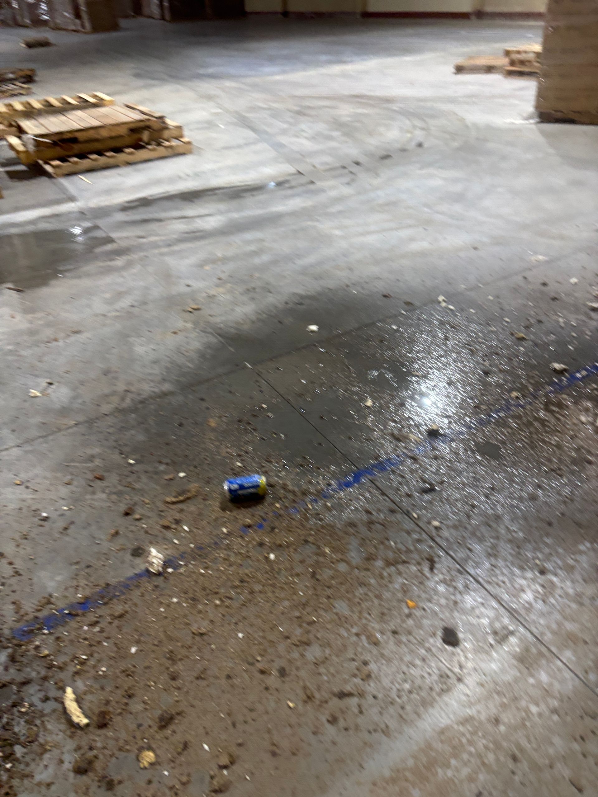 An empty warehouse floor with spilled substance and a discarded can. Pallets are stacked in the background.