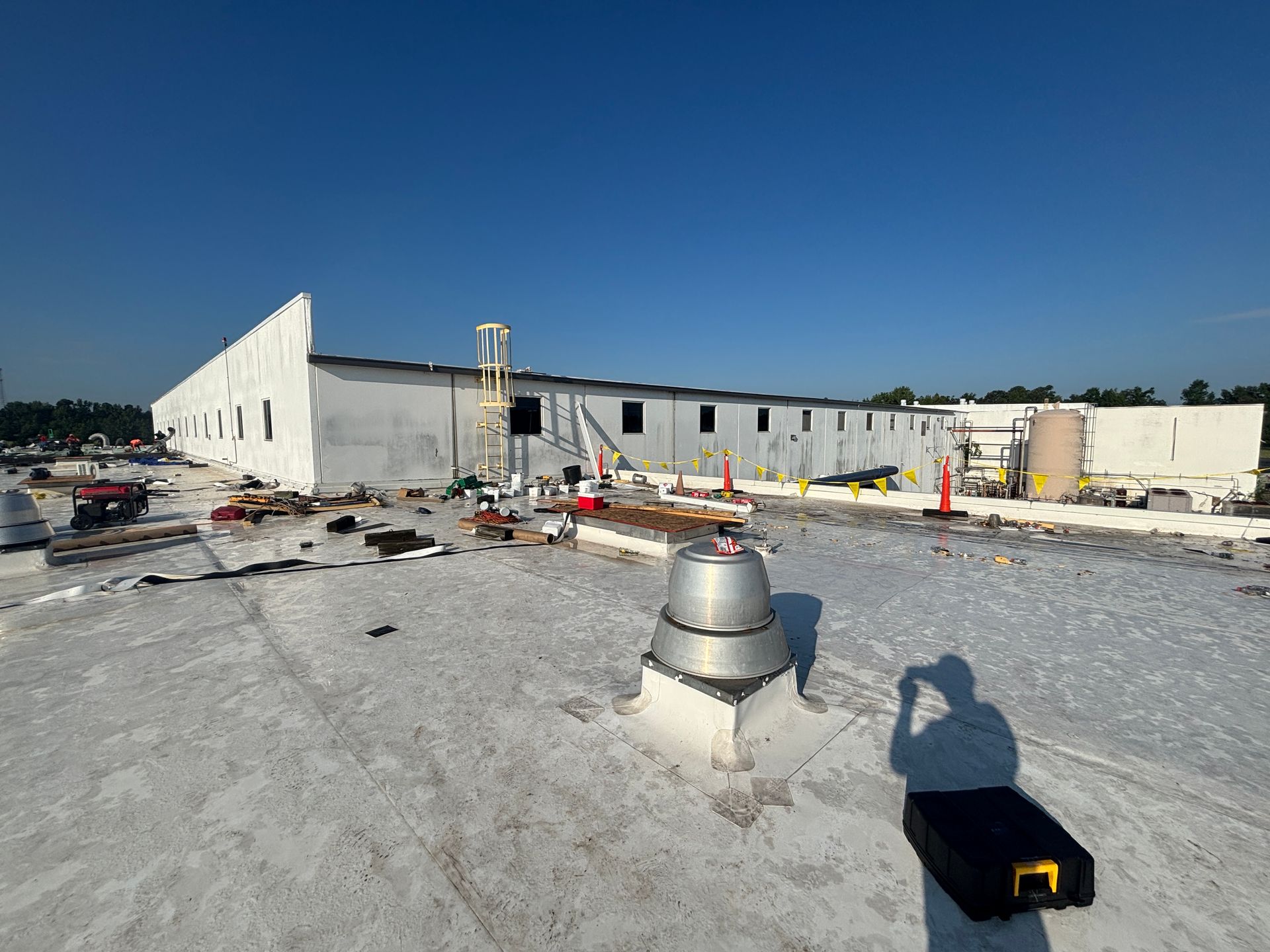 A white industrial building with ongoing construction. Blue sky overhead, worker's shadow in the foreground.