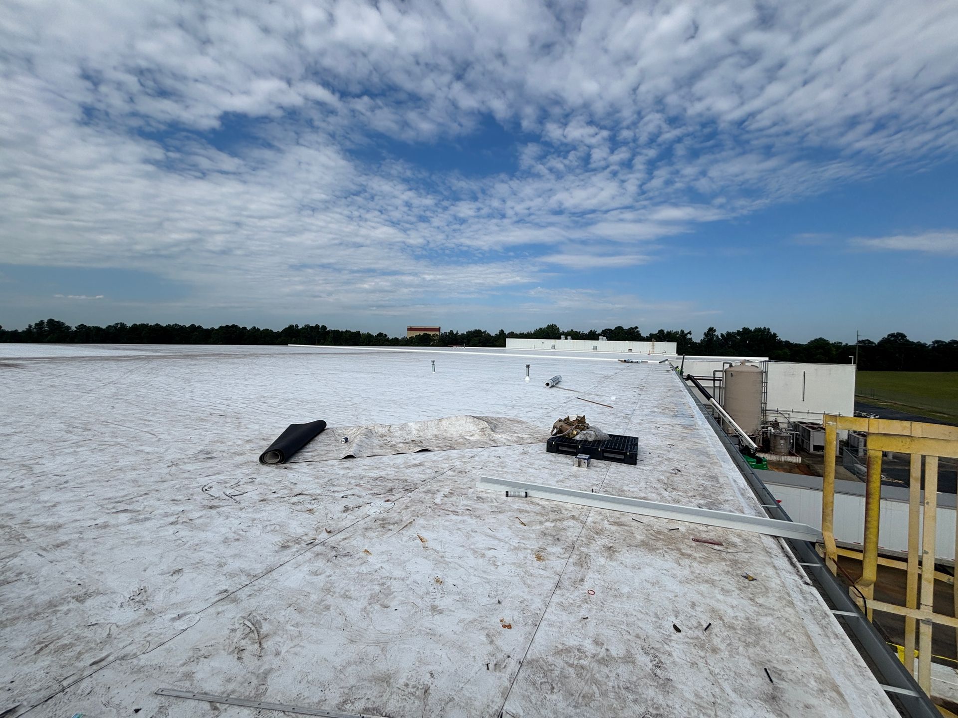 View of a large white flat roof on a partly cloudy day. Equipment and debris are visible on the roof.