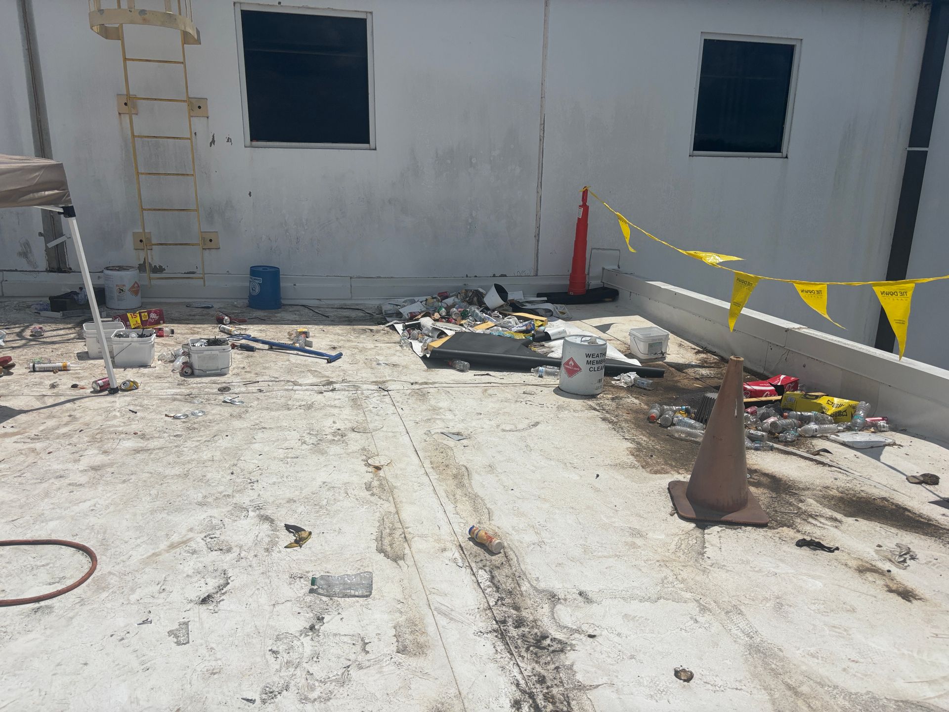 Rooftop with debris, ladder, safety cones, and caution tape. White building with two windows.