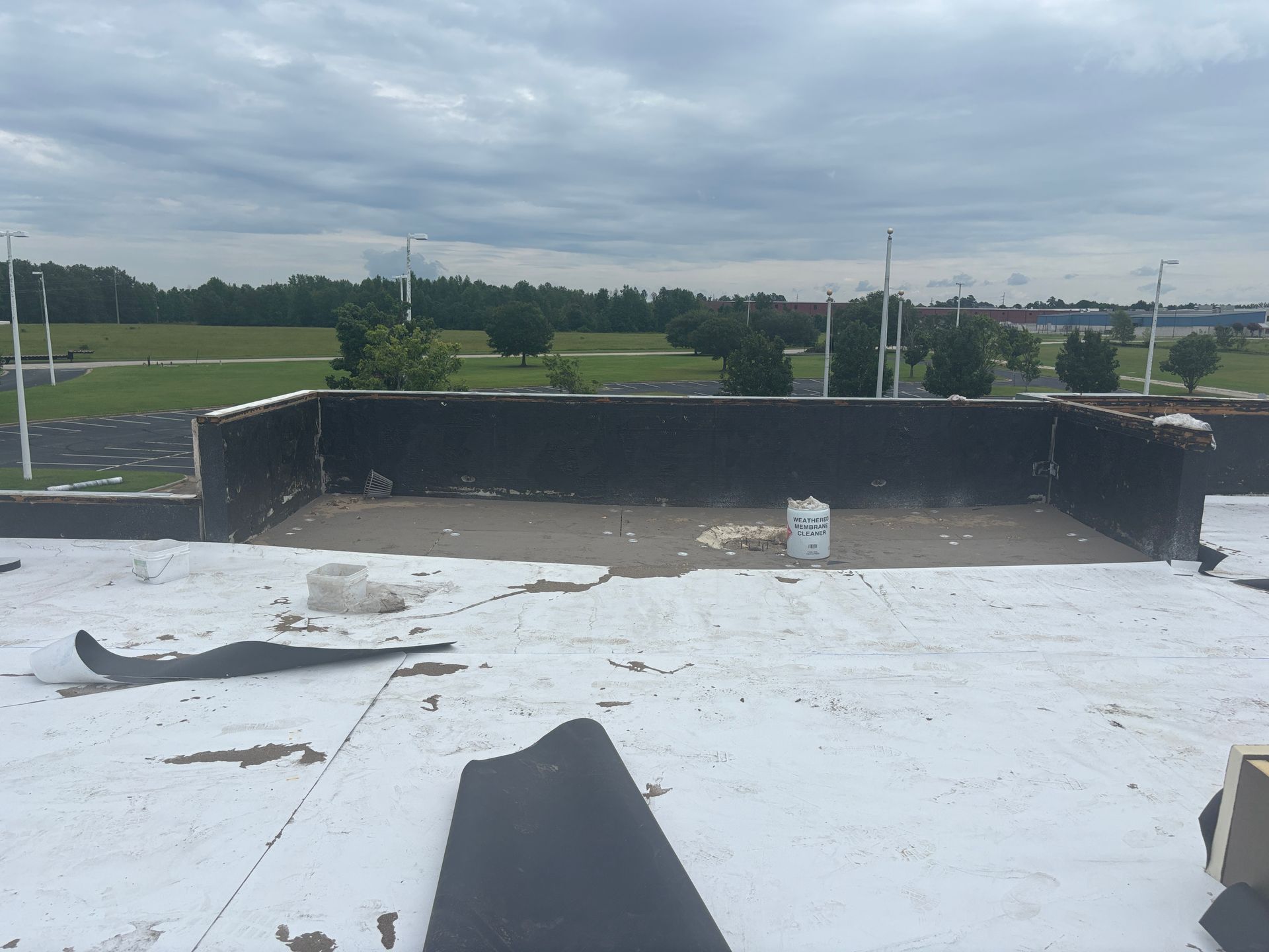 Rooftop with torn white membrane and black walls, looking towards green field under cloudy sky.