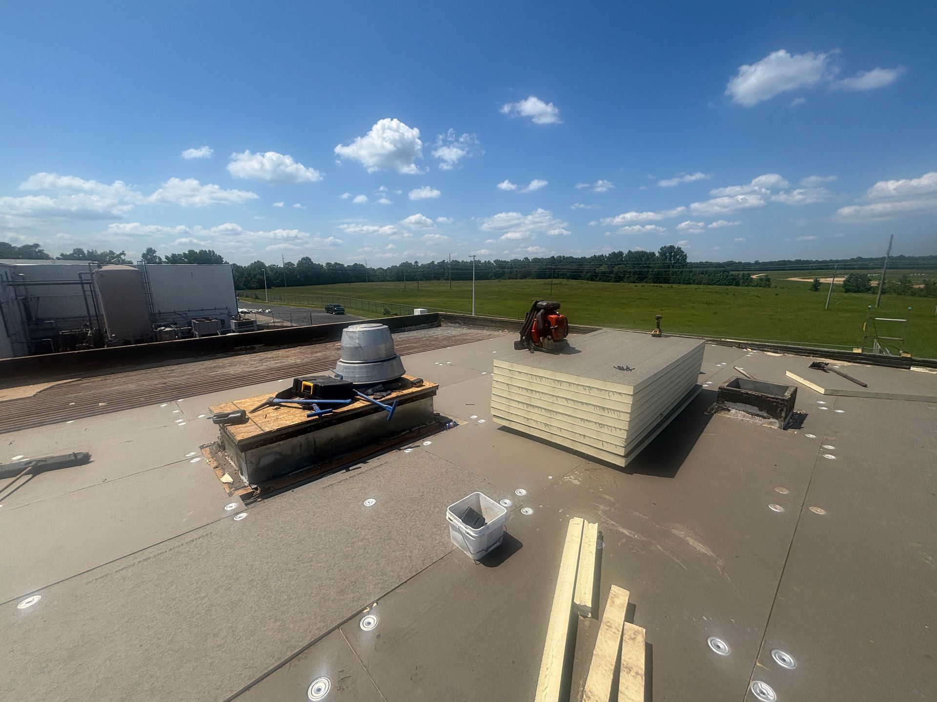Construction workers on a flat roof, placing insulation panels under a blue sky, surrounded by green fields.