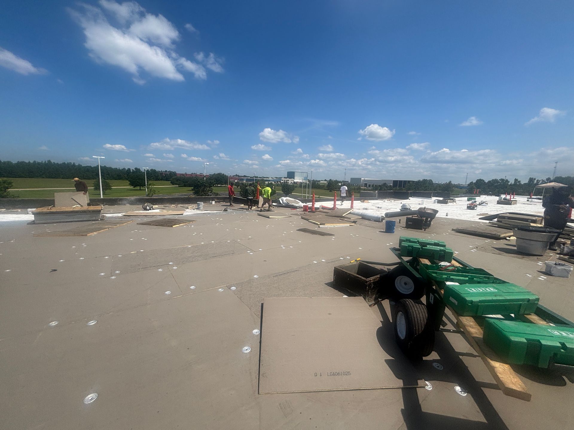 Workers on a flat roof under a blue sky; construction materials and equipment scattered.
