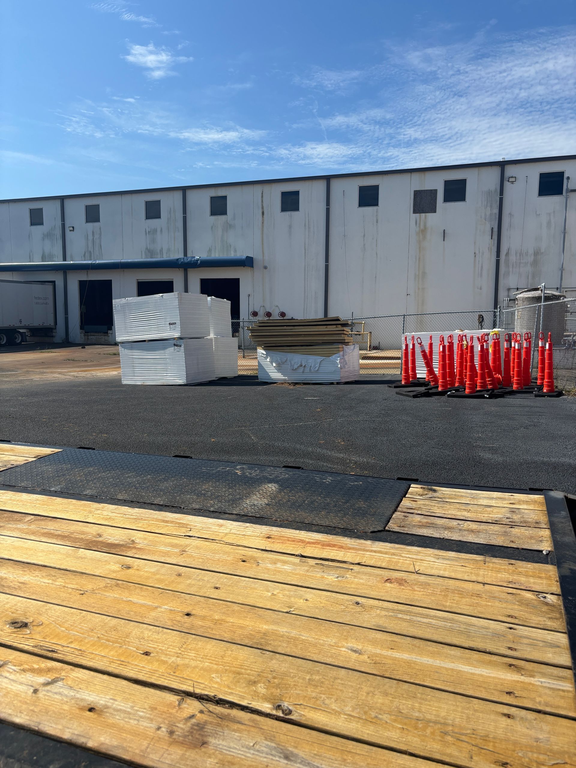 Exterior of a warehouse with construction materials. Wooden planks in foreground, dark gravel and cones. Blue sky overhead.