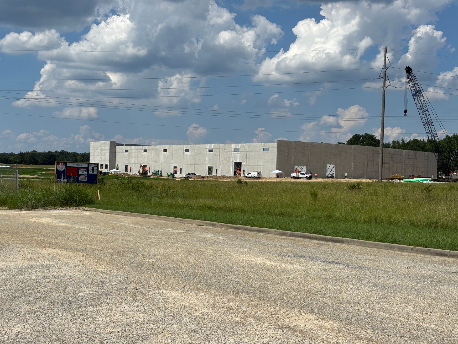 Large building under construction in a field, gray walls, blue sky with clouds, crane visible.