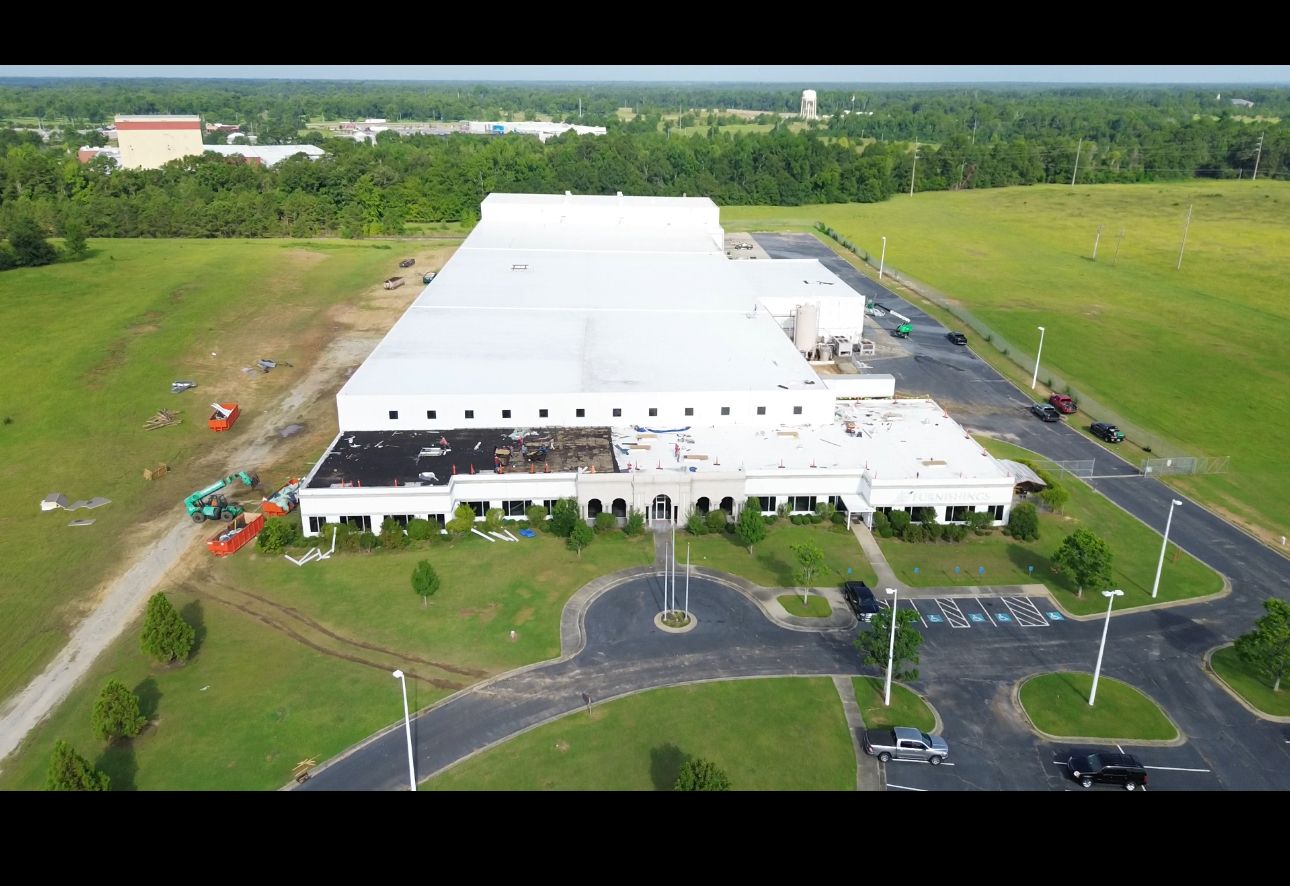 Aerial view of a large white industrial building with a flat roof, surrounded by green grass and a parking lot.